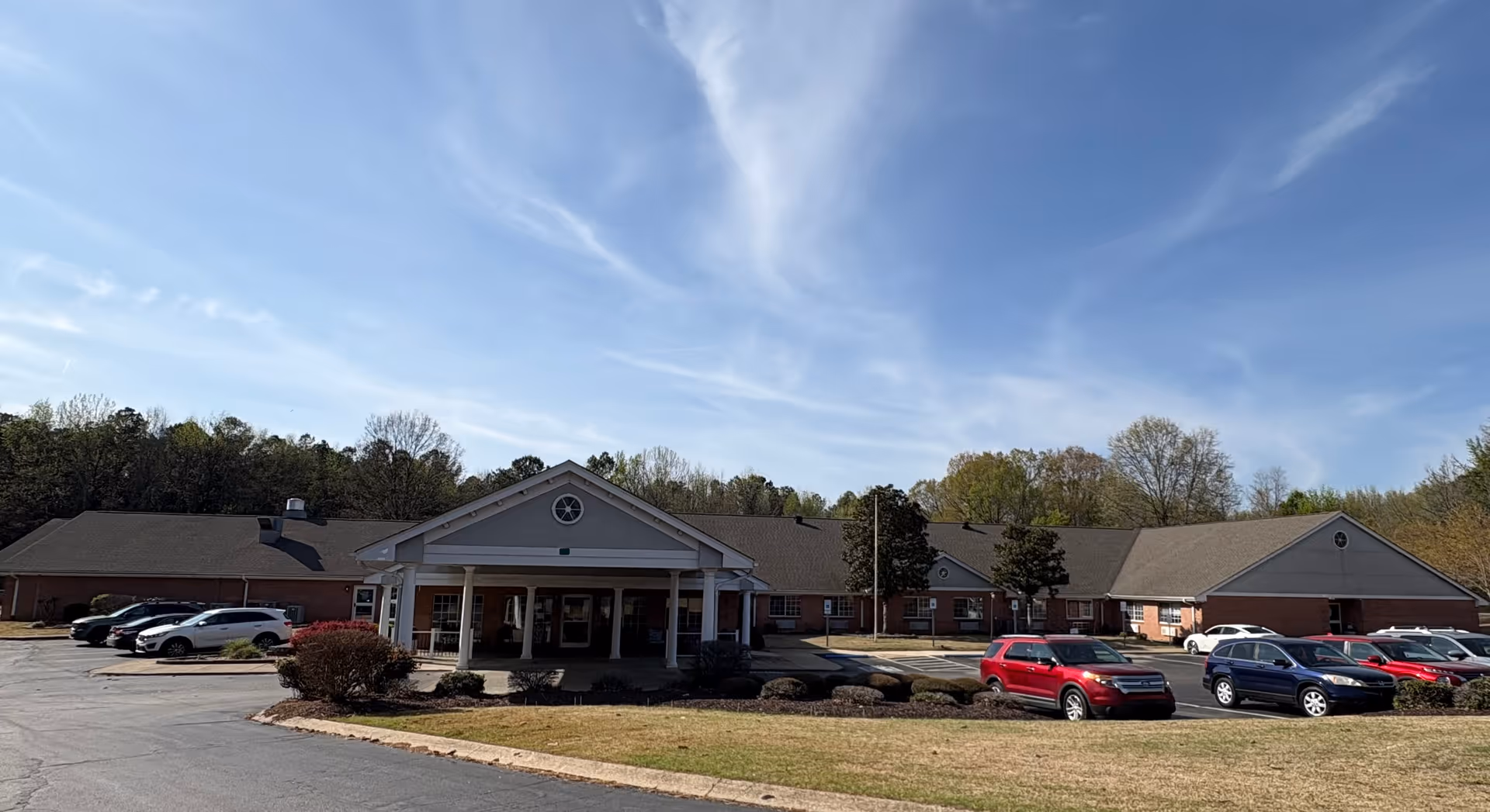 Single-story brick senior living building with a covered entrance, parked cars, lawn and trees under a blue sky.