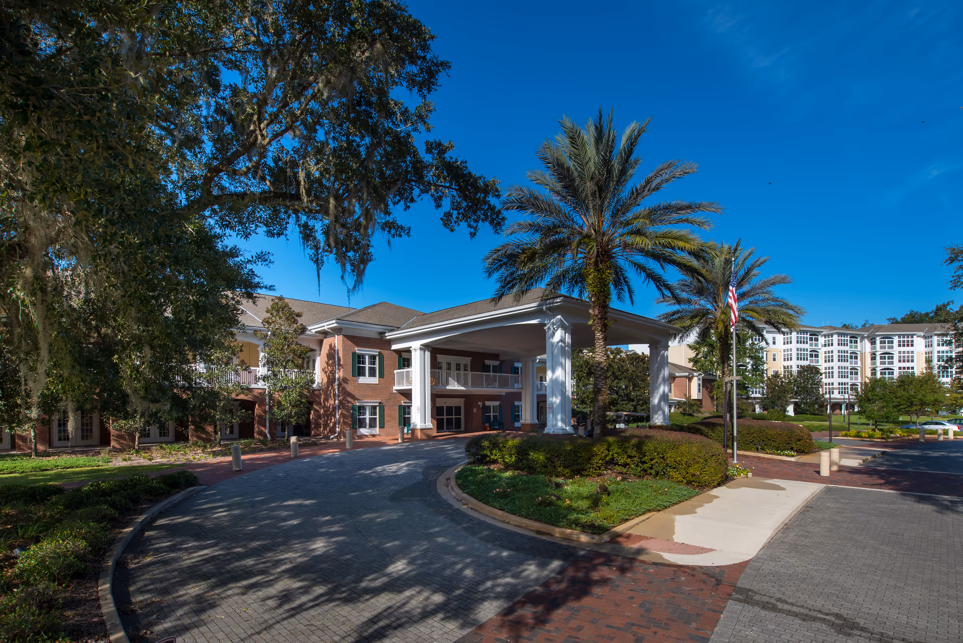 Entrance to a senior living facility with a covered drop-off area supported by white columns, surrounded by palm trees and other greenery under a clear blue sky. The building is a multi-story brick structure with white trim and balconies.