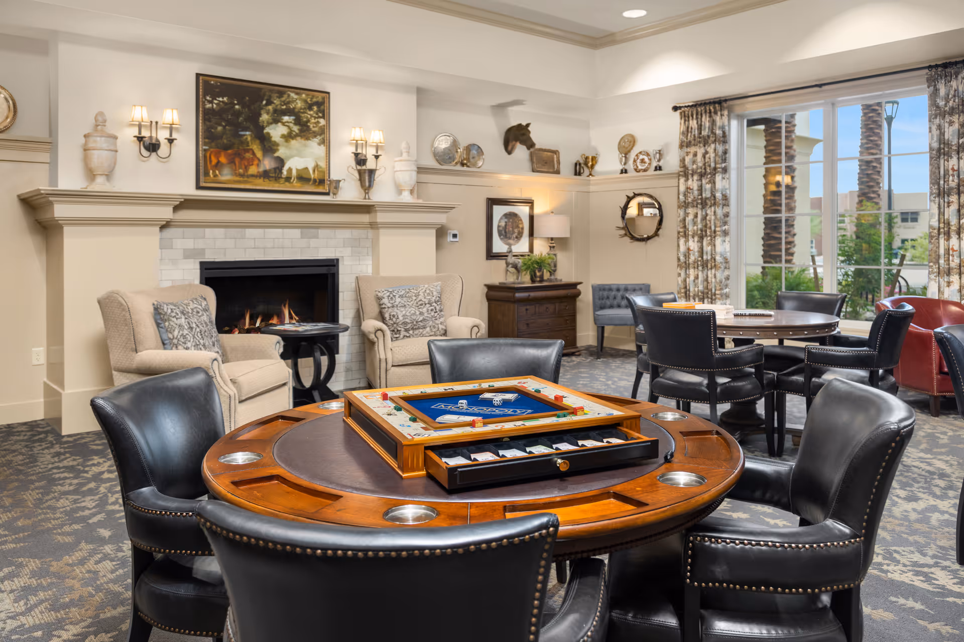 A cozy senior living community lounge with a round wooden game table featuring a board game and black leather chairs. In the background, there are two beige armchairs near a lit fireplace with a painting above it, decorative wall sconces, and a large window with patterned curtains letting in natural light. Additional tables and chairs are arranged around the room, with decorative items on shelves and walls.