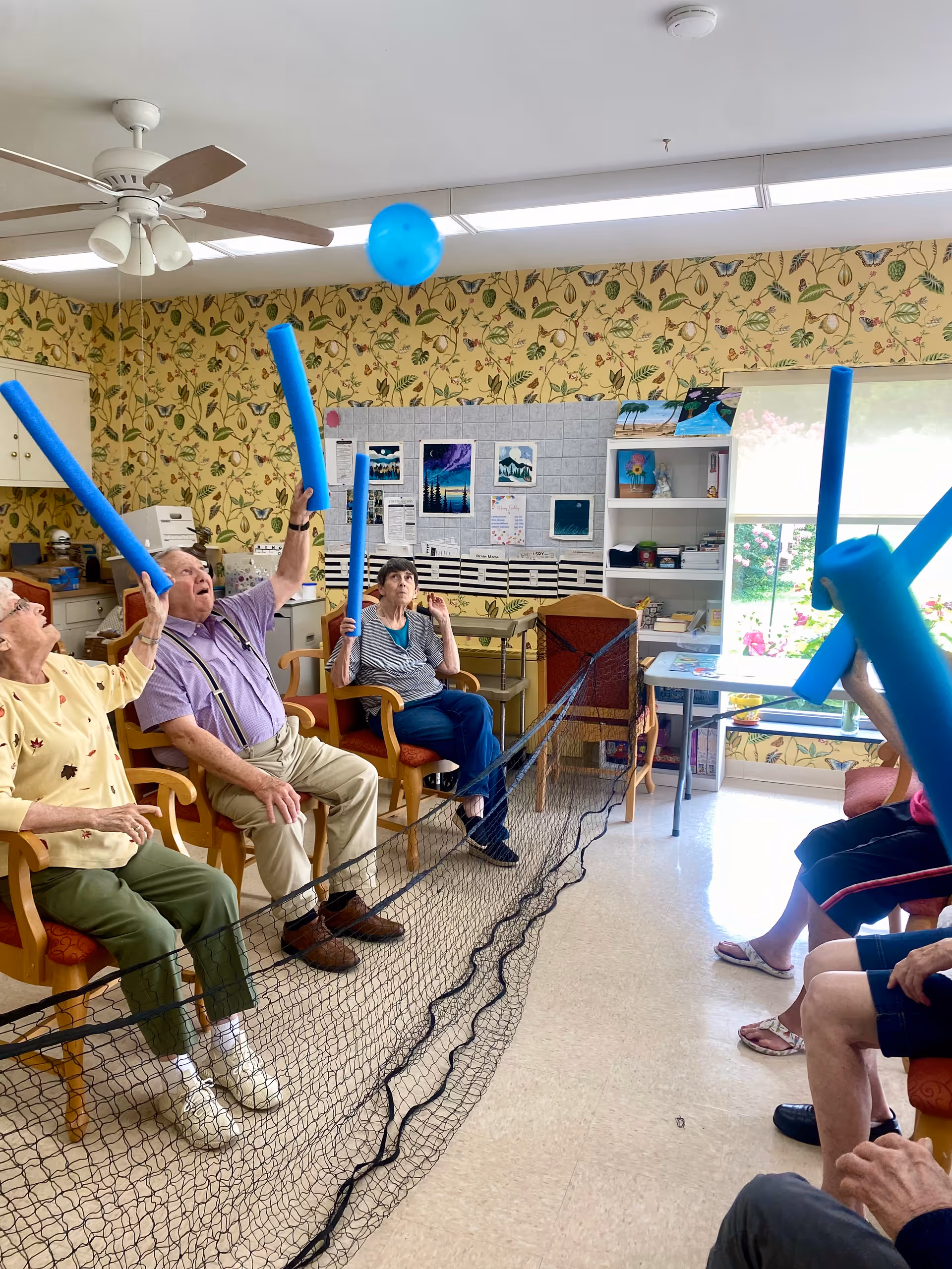 A group of elderly people sitting in chairs in a room with floral wallpaper, playing a seated balloon volleyball game using blue pool noodles to hit a blue balloon over a black net.