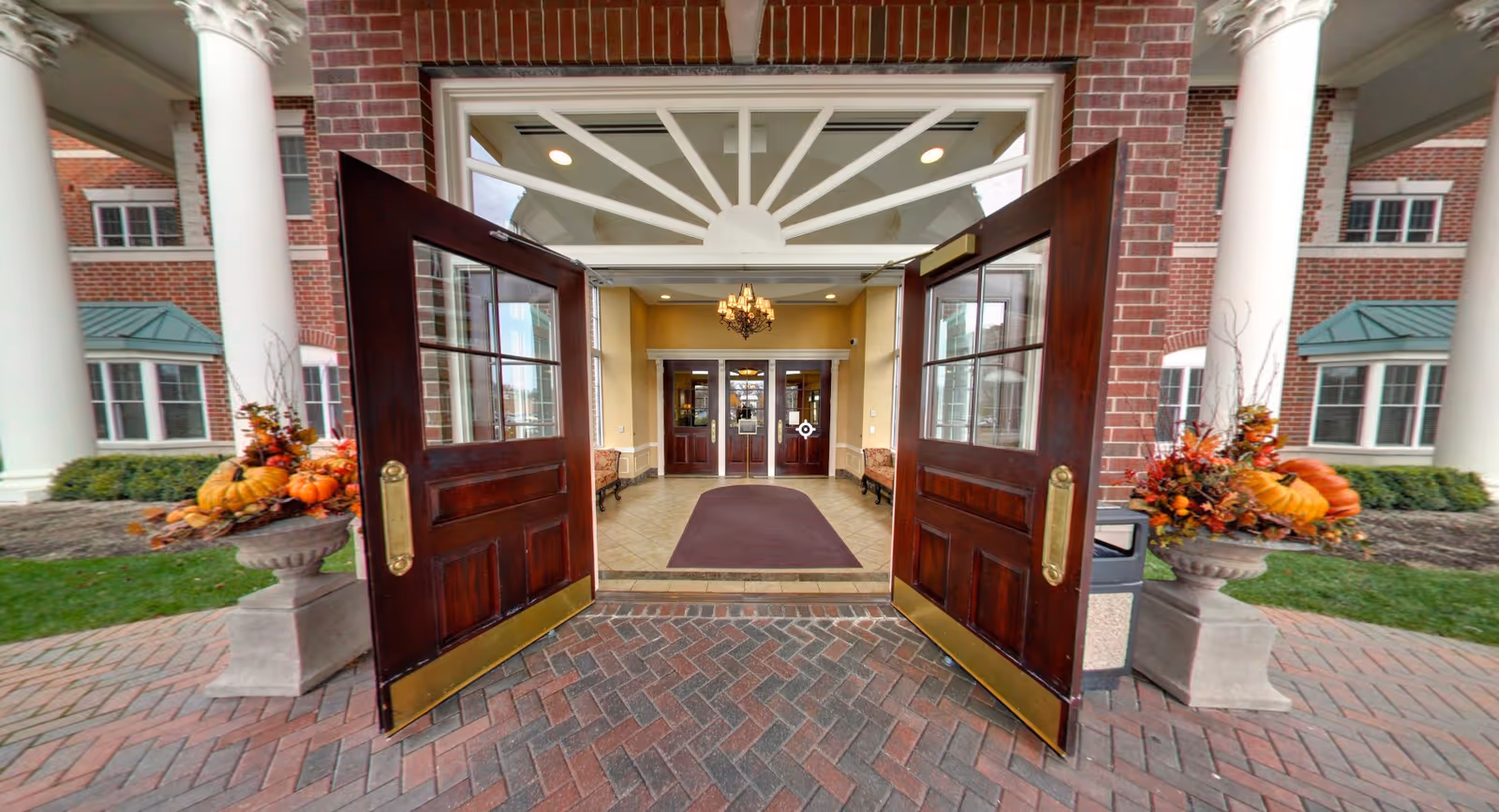 Open double wooden doors leading into the main entrance lobby of a brick building flanked by columns and pumpkin planters.