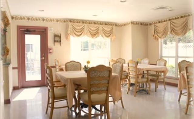 A bright dining room with multiple tables covered in beige tablecloths and surrounded by wooden chairs with cushioned seats. Large windows with beige valances allow natural light to fill the room. A door with glass panels is visible on the left side.