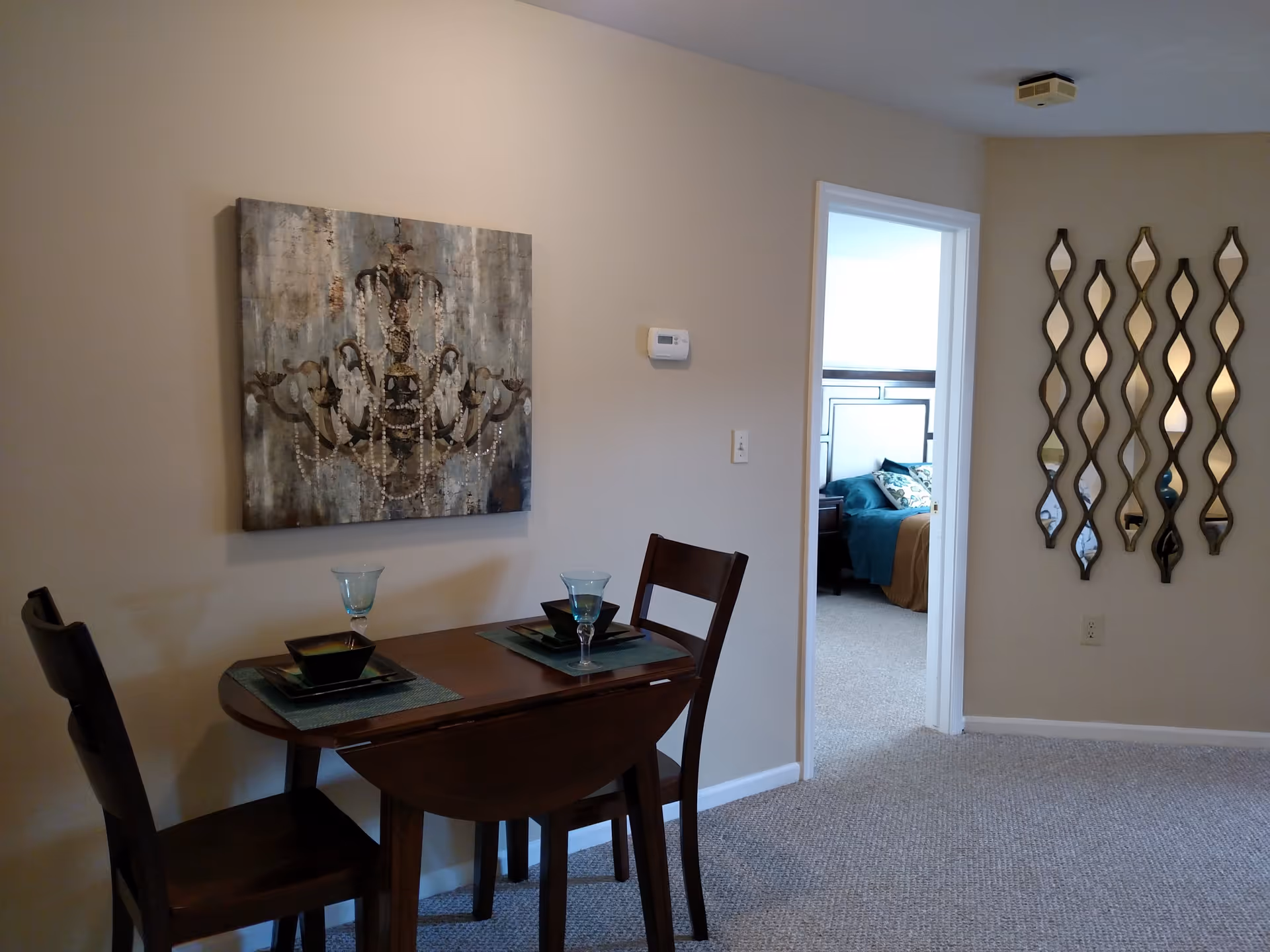 A small dining area with a wooden table set for two with blue placemats, bowls, and glasses. On the wall above the table is a painting of a chandelier. To the right, there is an open doorway leading to a bedroom with a bed covered in teal and brown bedding. On the adjacent wall, there are decorative wavy mirrors arranged vertically. The room has beige walls and carpeted flooring.