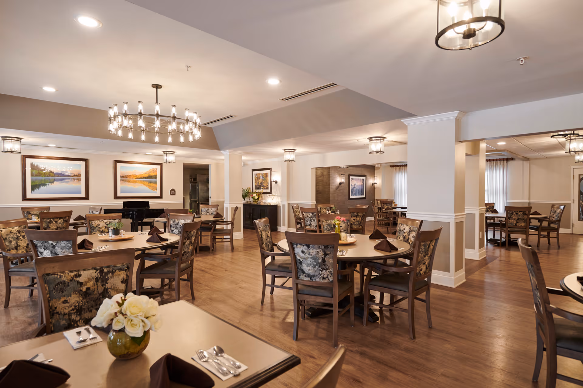 A spacious senior living dining room with multiple round tables and chairs arranged neatly. Each table is set with brown napkins, silverware, and small floral centerpieces. The room features warm wooden flooring, soft lighting from ceiling fixtures, and framed landscape paintings on the walls. Large windows with curtains allow natural light to enter the space.