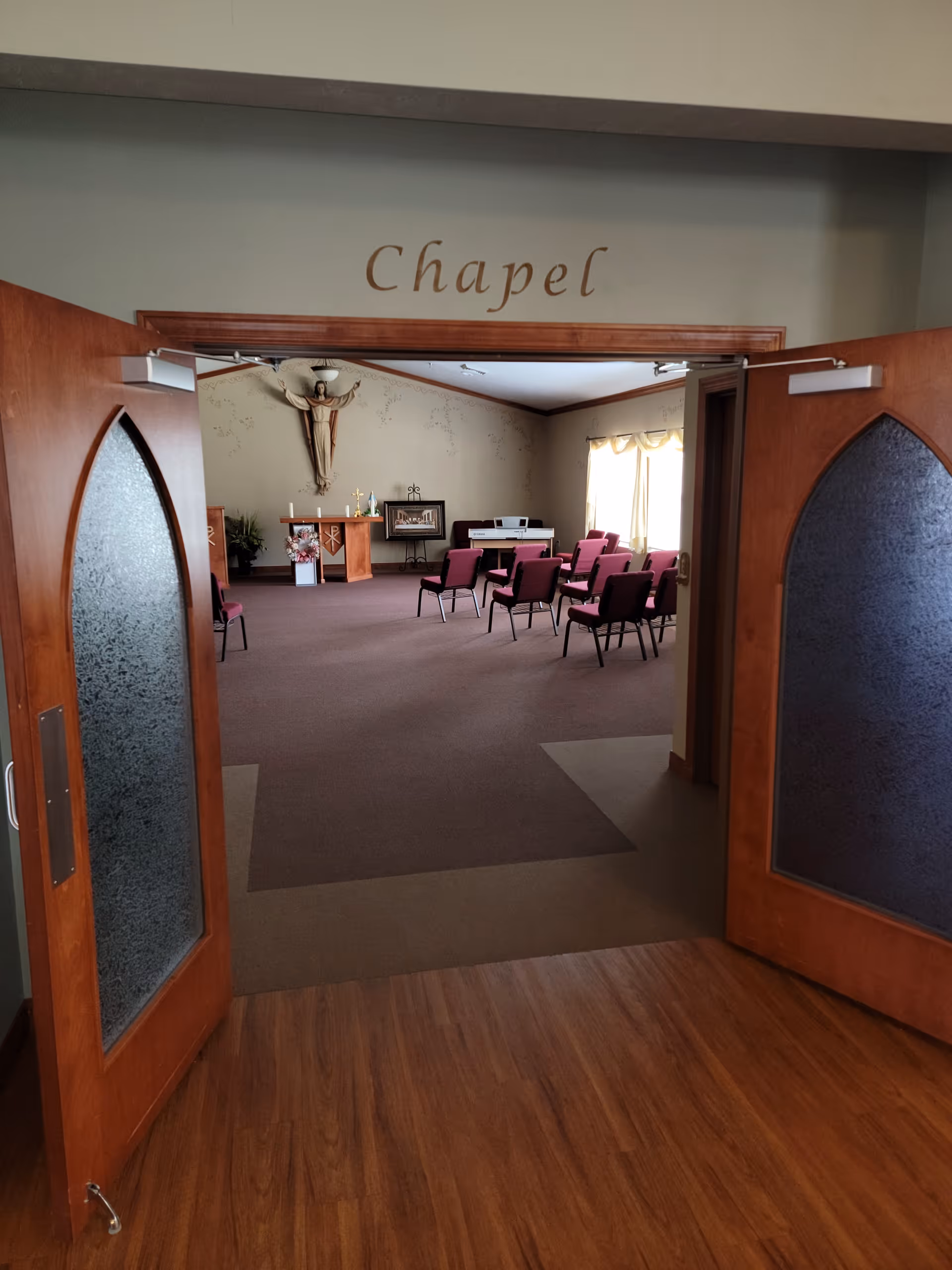 View through open wooden double doors into a chapel room with carpeted floor, rows of maroon chairs, a small altar with flowers and religious items, a wall-mounted crucifix, and a window with curtains letting in natural light.