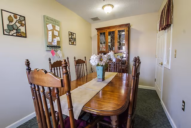 A dining room with a wooden dining table and six matching wooden chairs. A beige table runner and a vase with white flowers are placed on the table. On the far wall, there is a wooden china cabinet with glass doors. The walls are light-colored and decorated with two framed artworks and a bulletin board with notes. The room has a ceiling light fixture and a window with a valance on the right side.