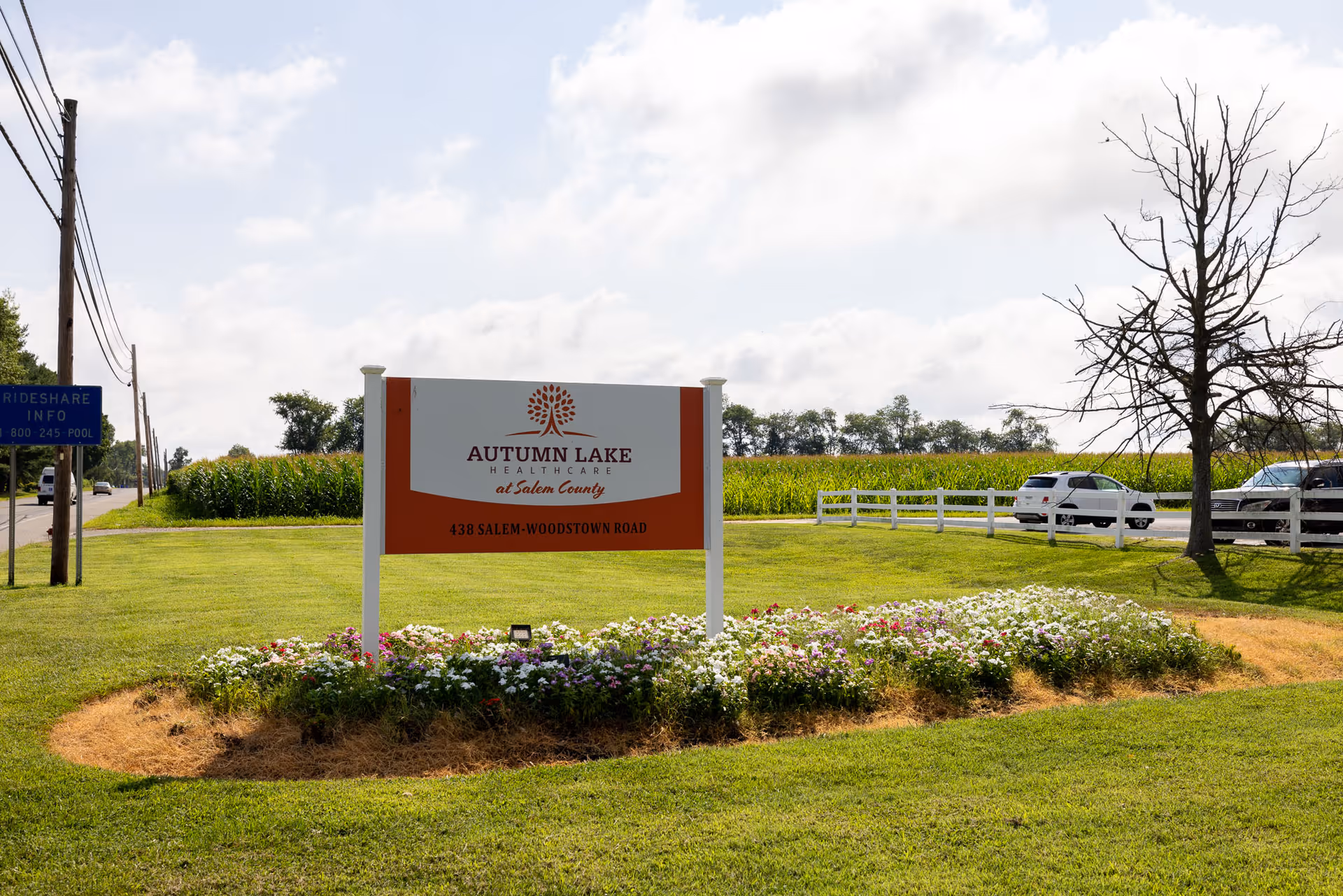 A sign reading "Autumn Lake Healthcare at Salem County" stands on a grassy lawn with flowers, a road and parked cars and fields in the background.