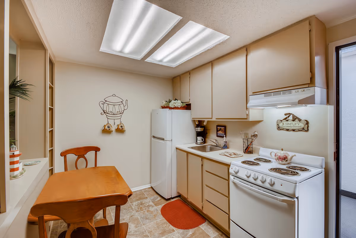 A small kitchen area with beige cabinets, a white refrigerator, a white electric stove with a floral teapot on top, a stainless steel sink, and a wooden table with a matching chair. The kitchen has tiled flooring and two fluorescent ceiling lights. There is a decorative wall hanging of a teapot with two mugs and a sign above the stove that says 'SIMPLIFY Live simply Live well'.