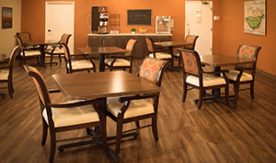 A dining room with several wooden tables and cushioned chairs arranged on a wood floor. The walls are painted a warm orange color, and there is a counter with a coffee machine and other items against the back wall. Two closed doors and framed artwork are visible on the walls.