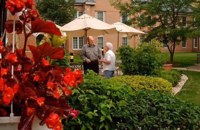 Two older adults talking in a landscaped courtyard with patio umbrellas and red flowers in the foreground.