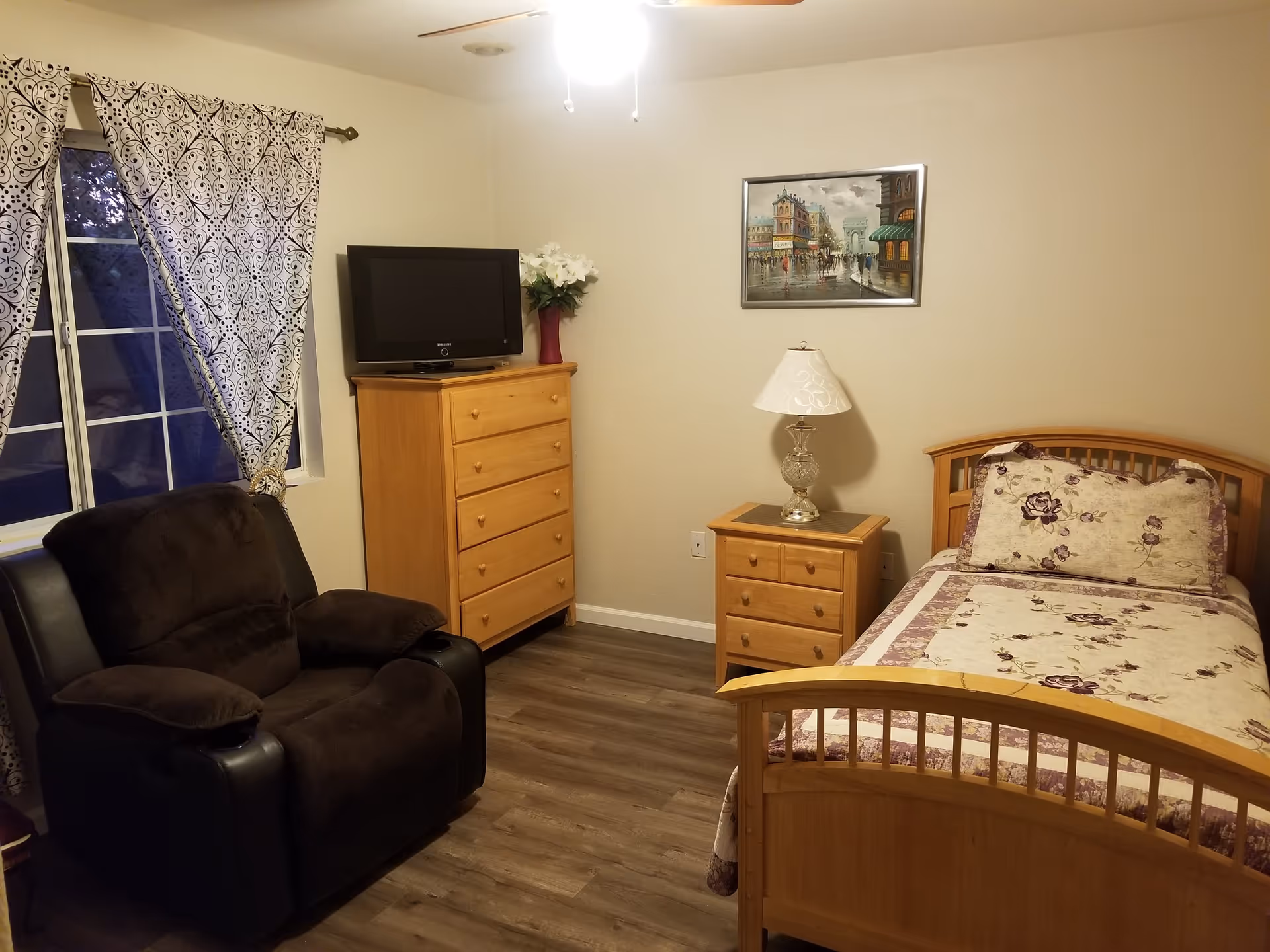 A cozy bedroom with a single wooden bed adorned with floral bedding and pillows. Next to the bed is a wooden nightstand with a decorative lamp. Across from the bed is a wooden dresser with a flat-screen TV on top and a vase with white flowers. A comfortable dark brown recliner chair is positioned near a window with patterned curtains. The room has wood flooring and a ceiling fan with a light.