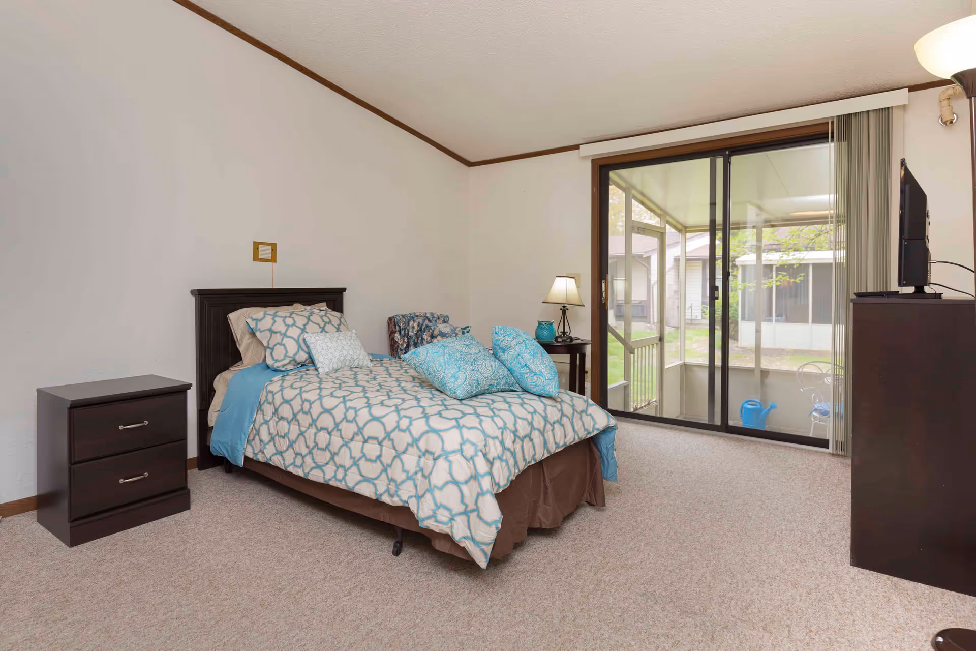 A bedroom with a single bed covered in a blue and white patterned comforter and pillows. There is a dark wooden nightstand with two drawers to the left of the bed and a small round table with a lamp and decorative vase to the right. A sliding glass door leads to an enclosed patio area with outdoor furniture and a watering can. A television sits on a dark wooden dresser on the right side of the room. The room has beige carpet and white walls with brown trim.