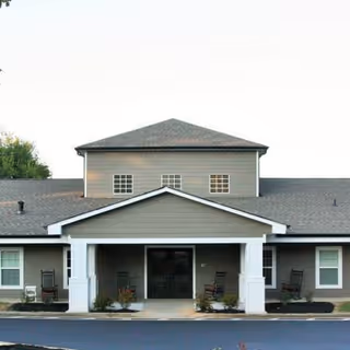 Front exterior view of a single-story senior living facility building with a covered entrance supported by white columns, several windows, and rocking chairs placed on the porch area.