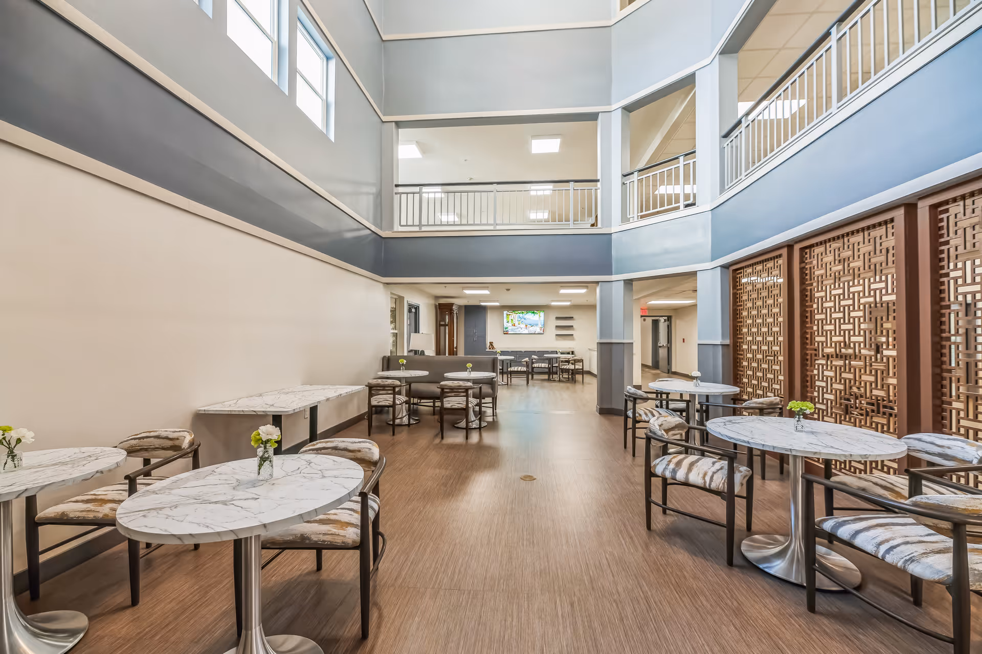 A bright and spacious indoor dining area with multiple round marble-top tables and cushioned chairs arranged neatly. The room features high ceilings with a second-floor balcony railing visible, large windows near the ceiling allowing natural light, and decorative wooden lattice panels on one side. There are small flower vases on each table, and a television mounted on the far wall above a seating area with additional tables and chairs.