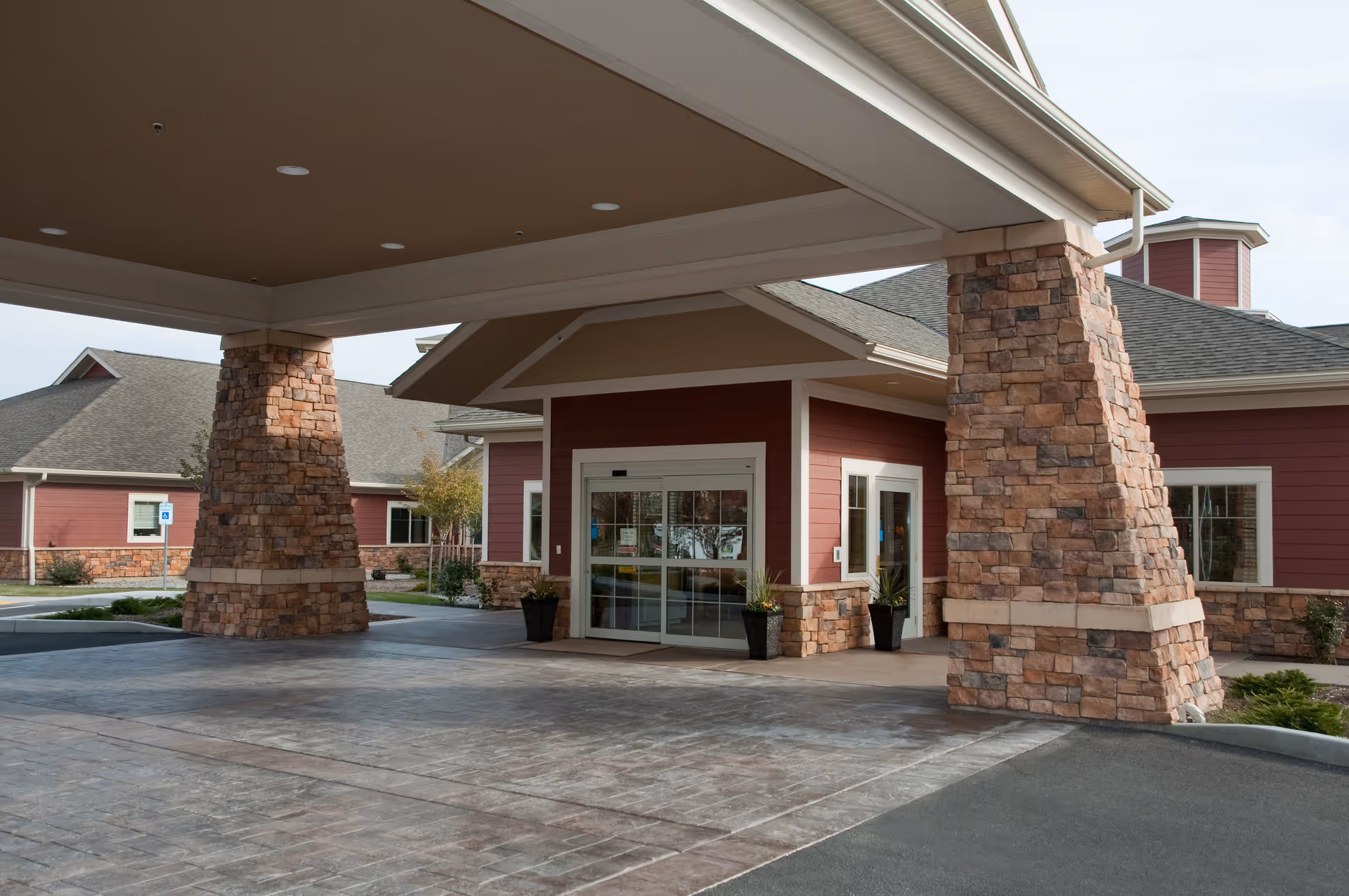 Entrance of a senior living facility with a covered drop-off area supported by large stone pillars. The building has red siding with white trim and stone accents, and automatic sliding glass doors at the entrance.