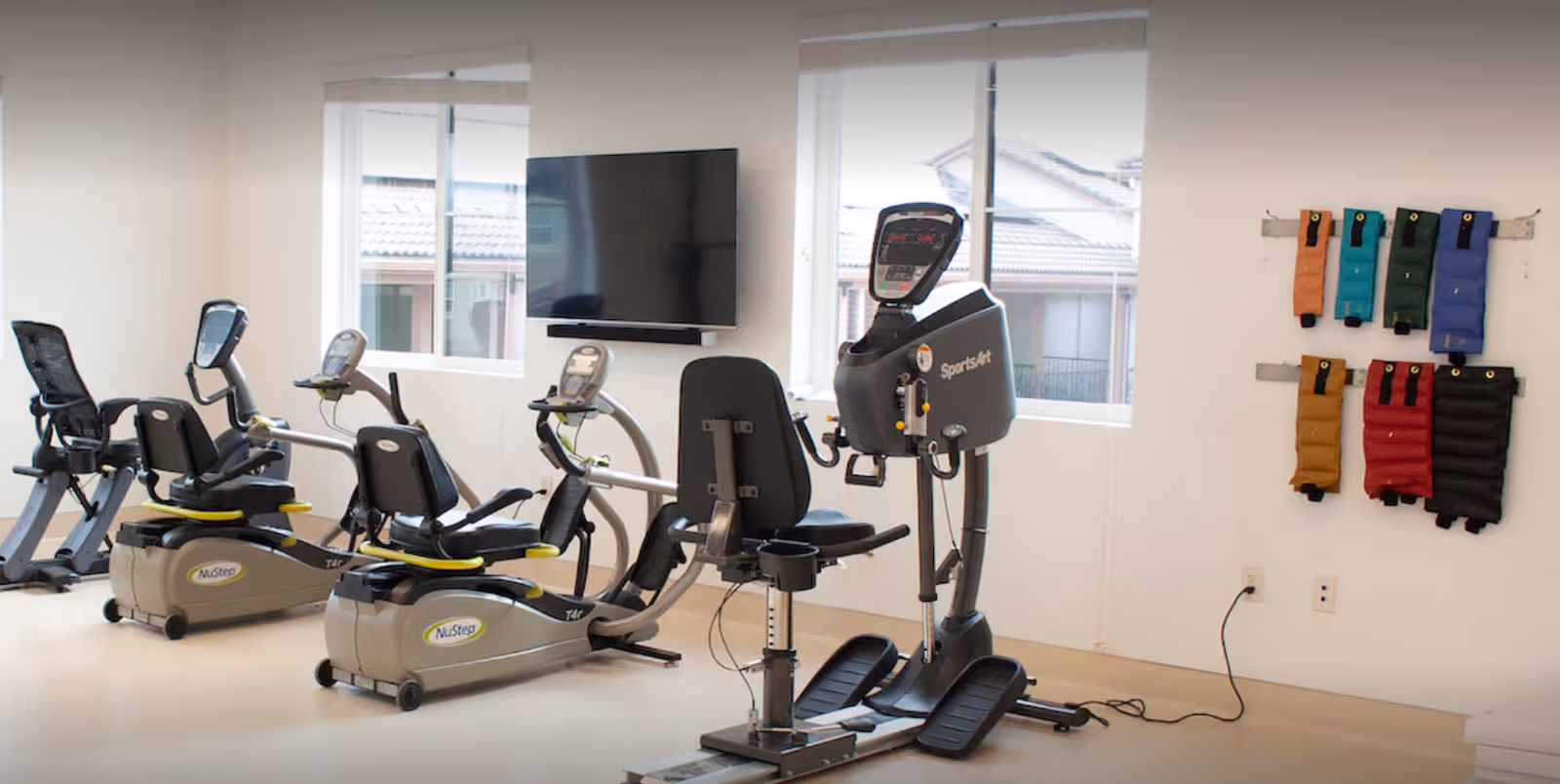 A fitness room with exercise equipment including three recumbent stationary bikes and one elliptical machine. There is a flat screen TV mounted on the wall between two windows. On the right wall, colorful ankle or wrist weights are hanging on racks.