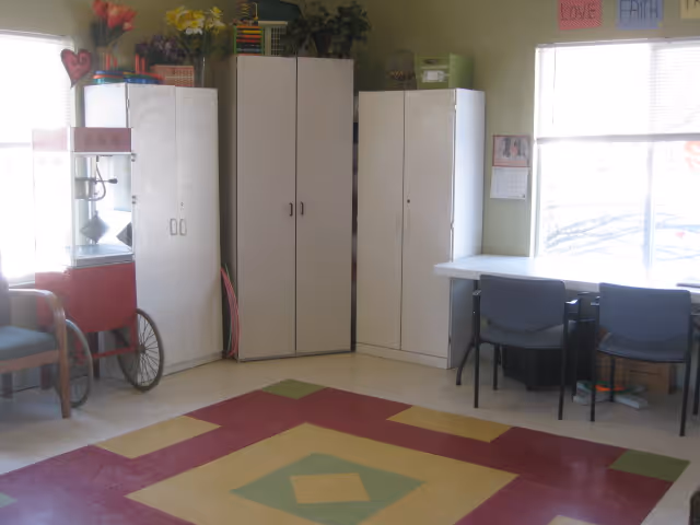 A room with a colorful geometric patterned floor, white storage cabinets against the wall, a popcorn machine on a cart, a table with two chairs near a window, and some decorative items including flowers and wall hangings.