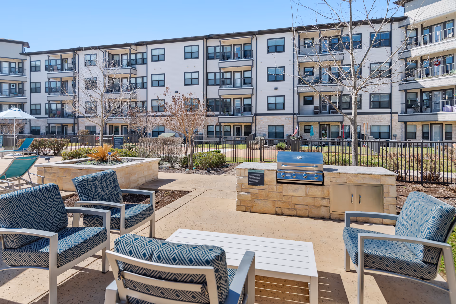 Outdoor patio area with cushioned chairs and a white table arranged around a built-in stone grill. In the background, there is a multi-story residential building with balconies and windows. The area is fenced and landscaped with small trees and shrubs under a clear blue sky.