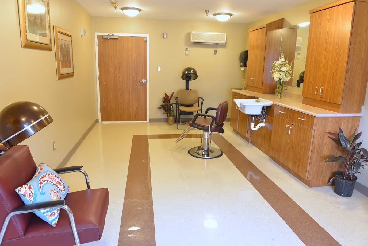 Interior view of a salon area in a senior living facility with a hair dryer chair, a styling chair, wooden cabinets with a sink, a vase with flowers, and two potted plants. The room has beige walls, a wooden door, and framed artwork on the wall.