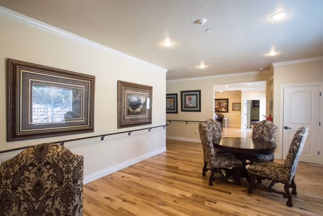 A well-lit interior room with wooden flooring, beige walls, and decorative framed artwork on the walls. There is a round wooden table with four upholstered chairs featuring a floral pattern. The room has ceiling lights and a handrail along the wall.