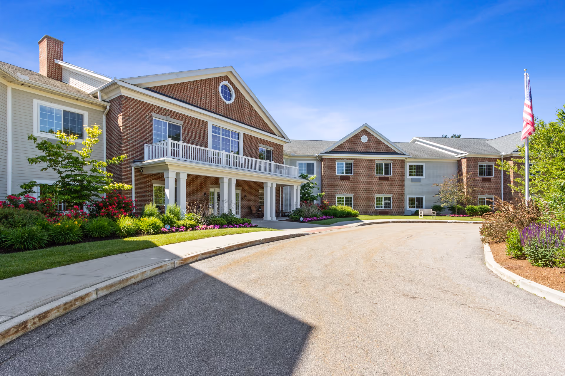 Front exterior view of a two-story senior living facility building with a brick facade and white siding. The entrance features a covered porch with white columns and a balcony above. The surrounding area includes well-maintained landscaping with green shrubs, colorful flowers, and trees. An American flag is visible on a flagpole to the right, under a clear blue sky.