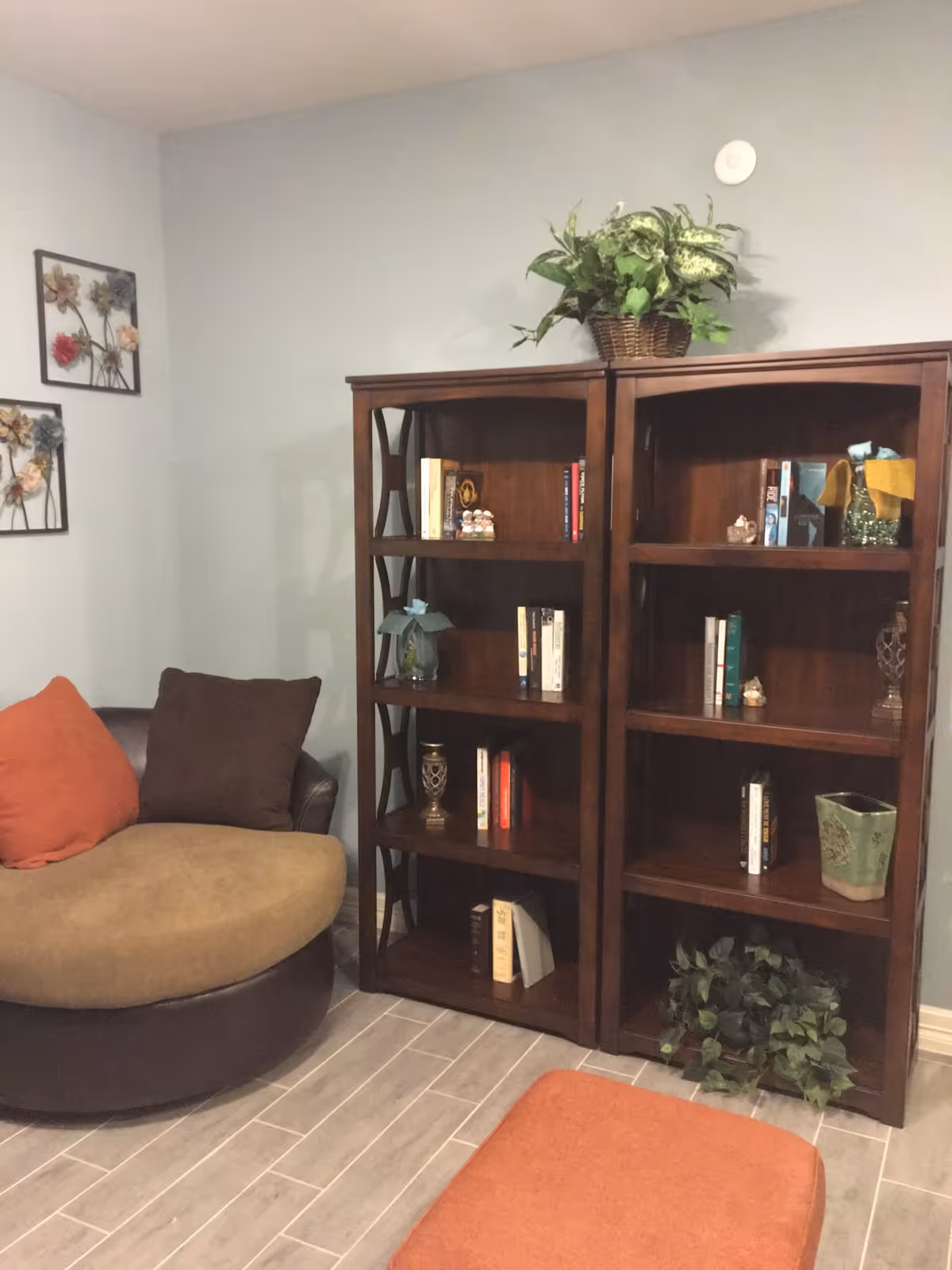 Cozy corner of a living room with a round cushioned chair featuring orange and brown pillows, two wooden bookshelves filled with books and decorative items, a green plant on top of the shelves, and a small orange ottoman on a tiled floor.