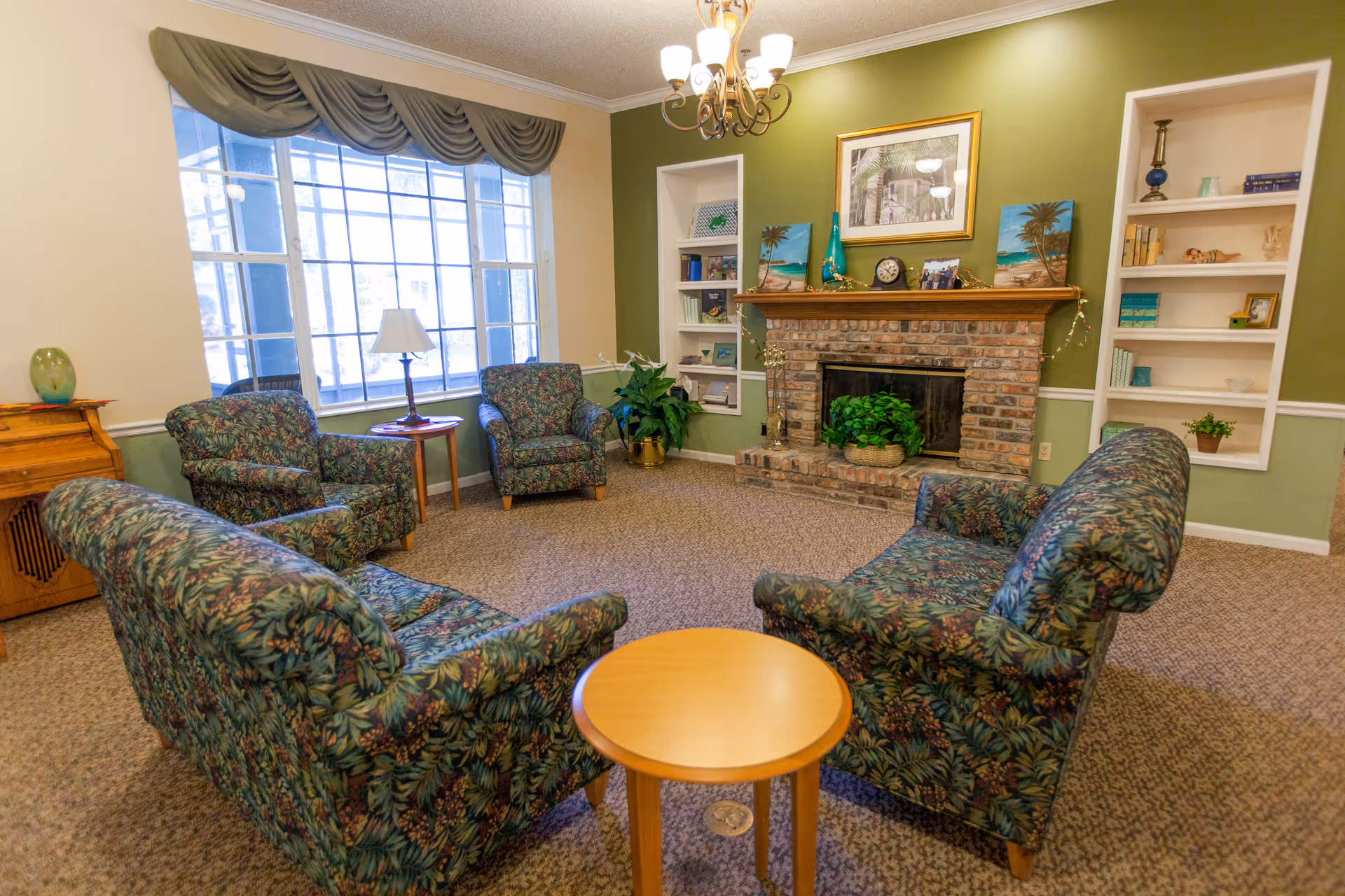 A cozy living room with floral patterned sofas and armchairs arranged around a small wooden round table. The room features a brick fireplace with a wooden mantle decorated with framed pictures, a clock, and small decorative items. Built-in white shelves flank the fireplace, holding books and decorative objects. A large window with green drapes allows natural light to fill the room, and a chandelier hangs from the ceiling.