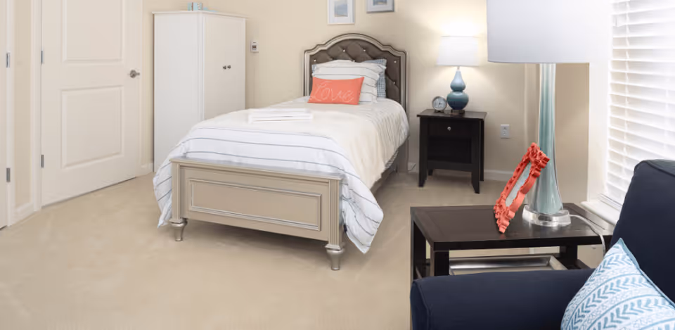 A neatly arranged bedroom featuring a single bed with white and gray striped bedding and a coral pillow with the word 'Love'. Next to the bed is a black nightstand with a blue lamp and a small clock. A white cabinet stands against the wall near a closed door. In the foreground, there is a dark blue armchair with a patterned cushion and a black side table holding a tall blue vase and a red decorative frame. The room has beige walls and carpet, with a window covered by white blinds.