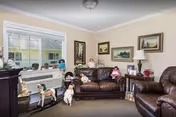 A cozy living room with brown leather sofas and armchairs, decorated with various dog figurines and paintings on the walls. There is a large window letting in natural light and a small side table with a lamp and decorative items.