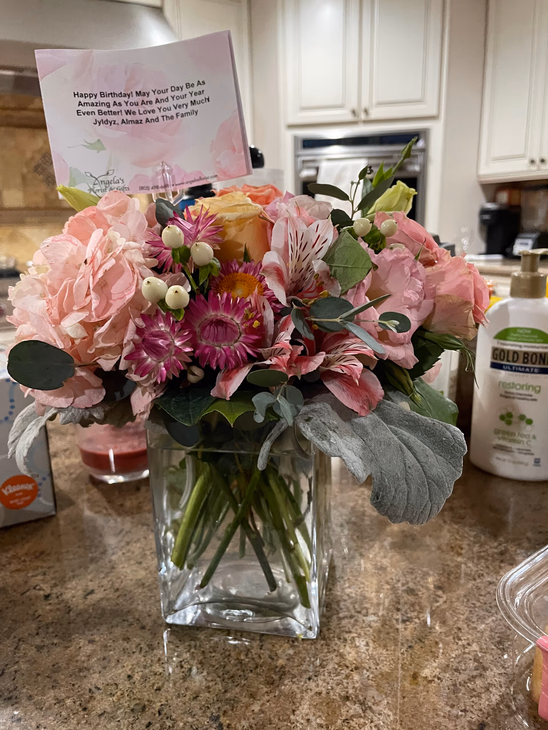A colorful bouquet of flowers in a clear square glass vase placed on a kitchen countertop. The bouquet includes pink, purple, and yellow flowers with green leaves. A birthday card is inserted in the bouquet with a message. In the background, there are white kitchen cabinets, a built-in oven, a coffee maker, a bottle of Gold Bond lotion, and a box of Kleenex tissues.