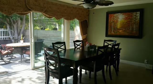 Sunlit dining room with a dark wooden table and chairs, a ceiling fan, wall art, and sliding glass doors opening to a patio with outdoor seating.