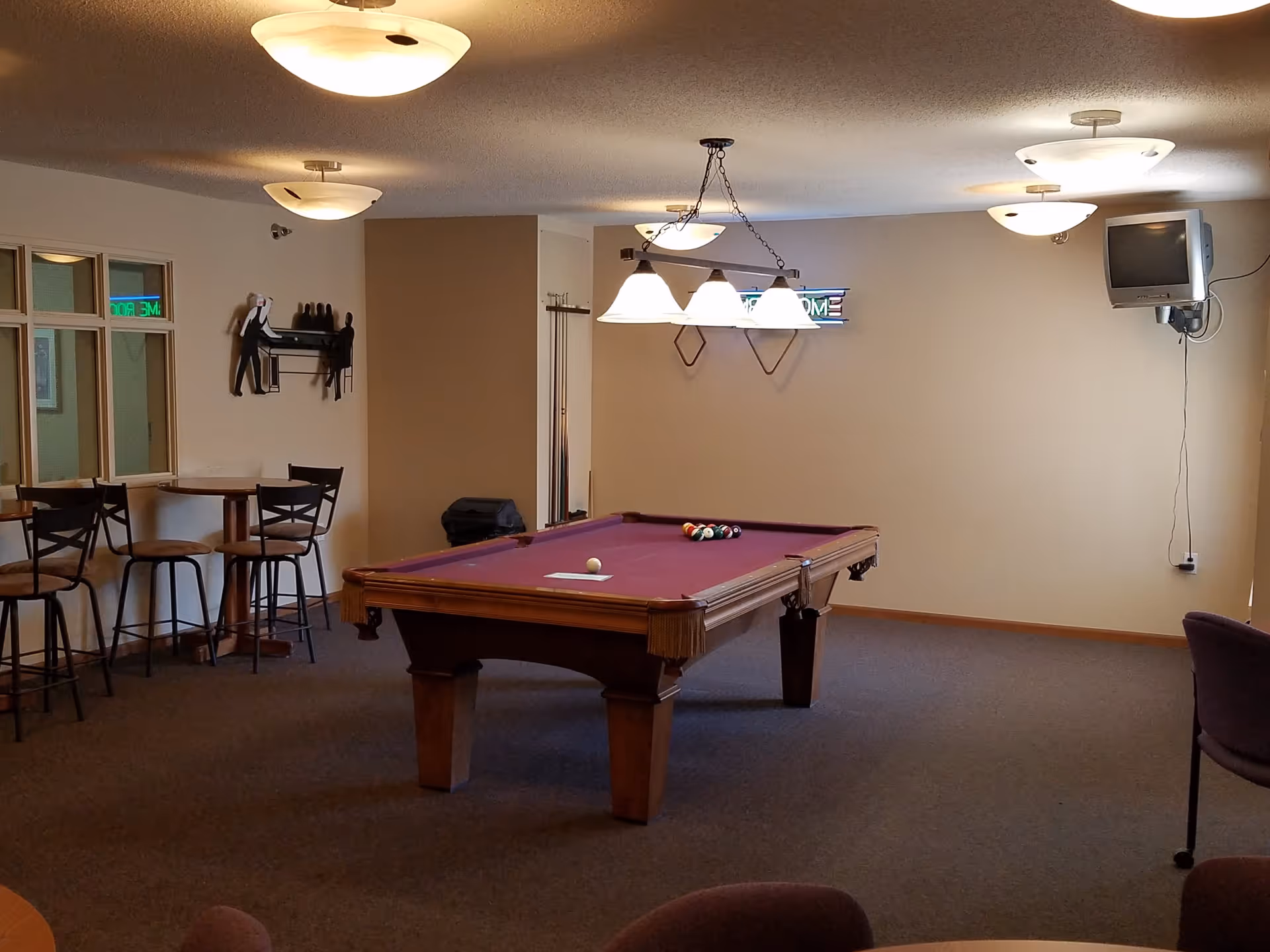 A recreational room with a pool table in the center, a set of pool balls arranged on the table, overhead lights, a small wall-mounted TV, a neon sign that says 'GAME ROOM', and a few tables with chairs along the wall.