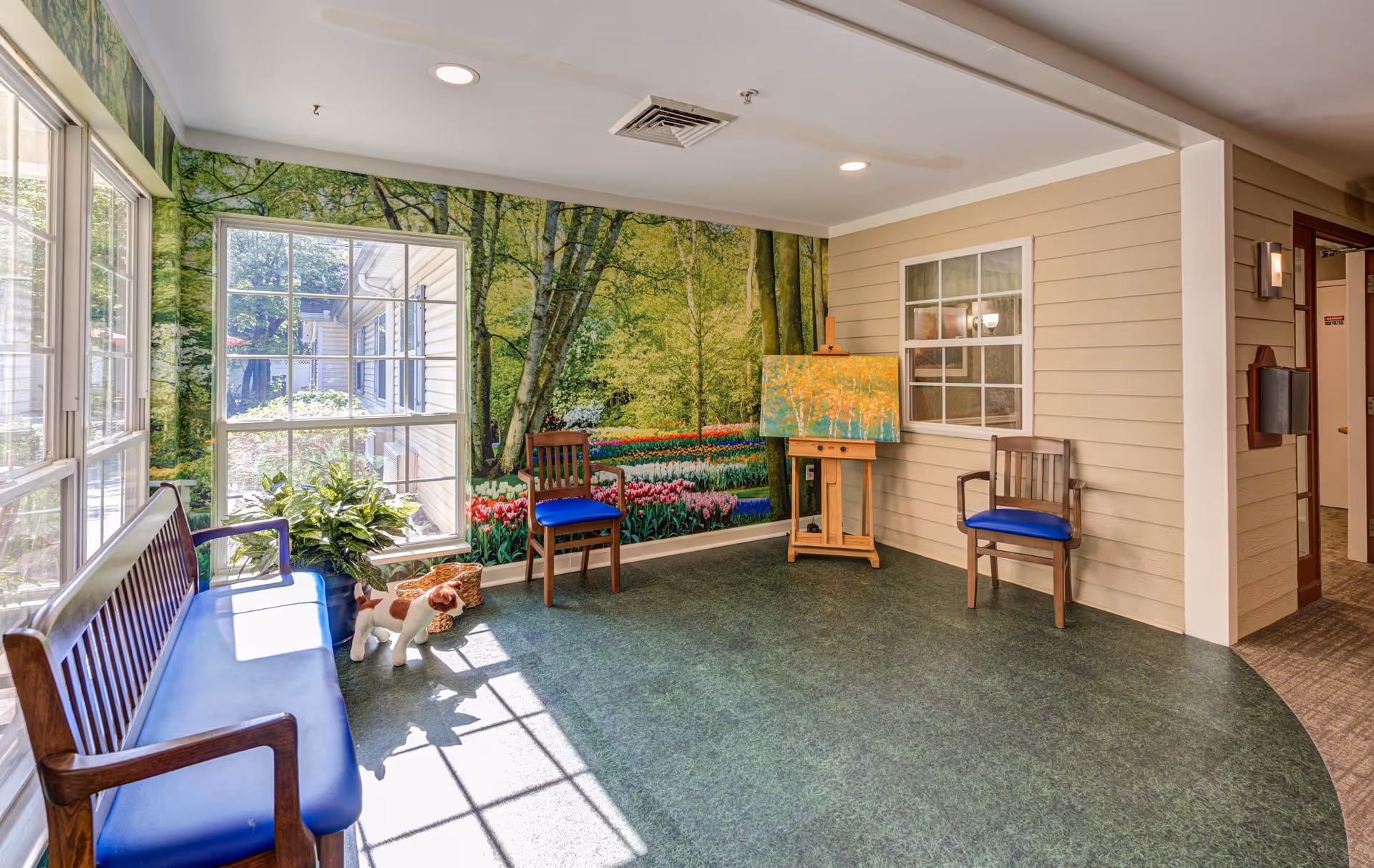A bright indoor seating area with large windows letting in natural light. The room has green flooring and beige walls, with a mural of a forest and tulip garden on one wall. There are three wooden chairs with blue cushions and a wooden bench with a blue cushion. A small dog statue and a potted plant are near the window. An easel with a colorful painting is positioned against the wall.