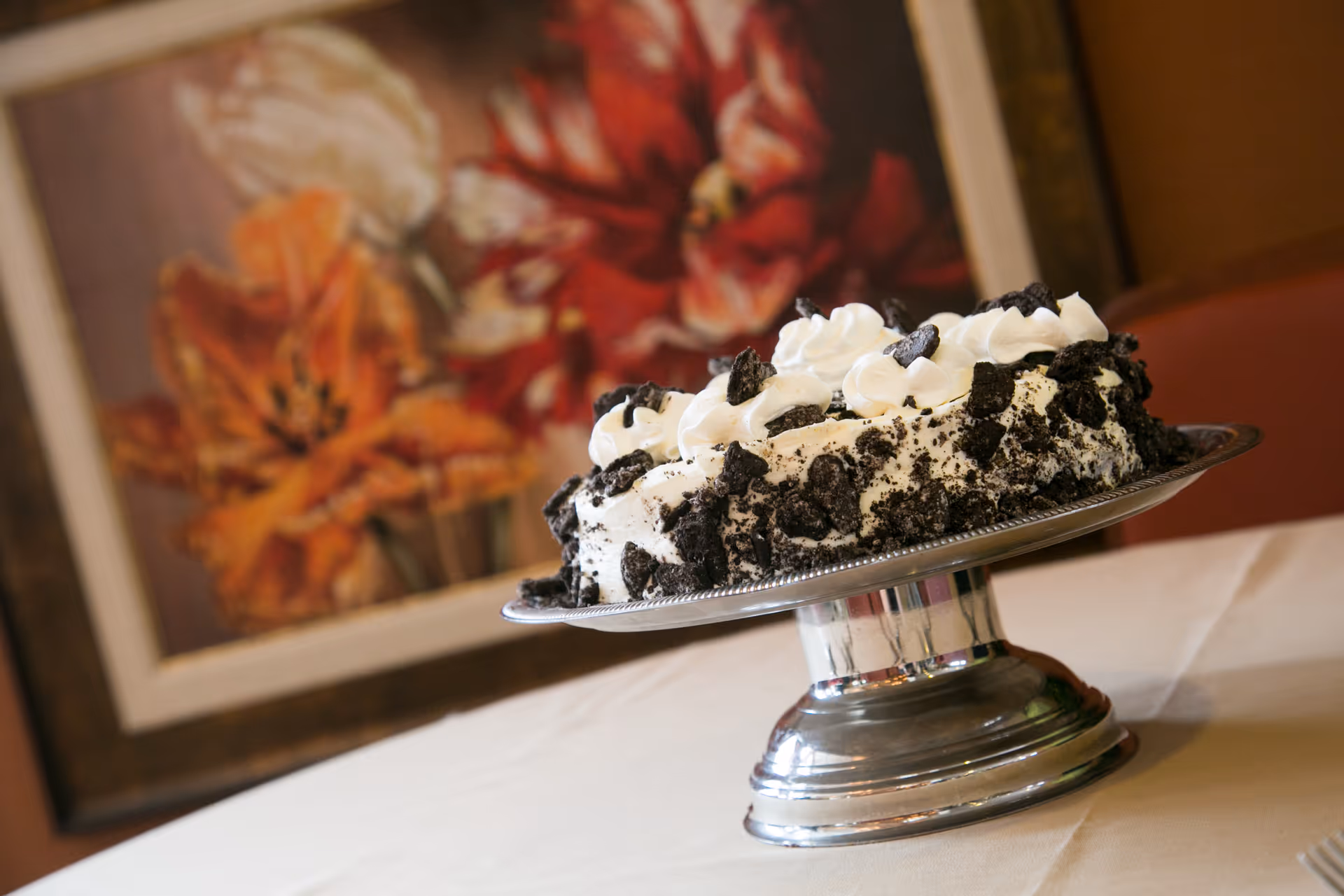 A cookies and cream cake with whipped cream dollops on top, placed on a silver cake stand on a white tablecloth. In the background, there is a framed painting of orange and red flowers.