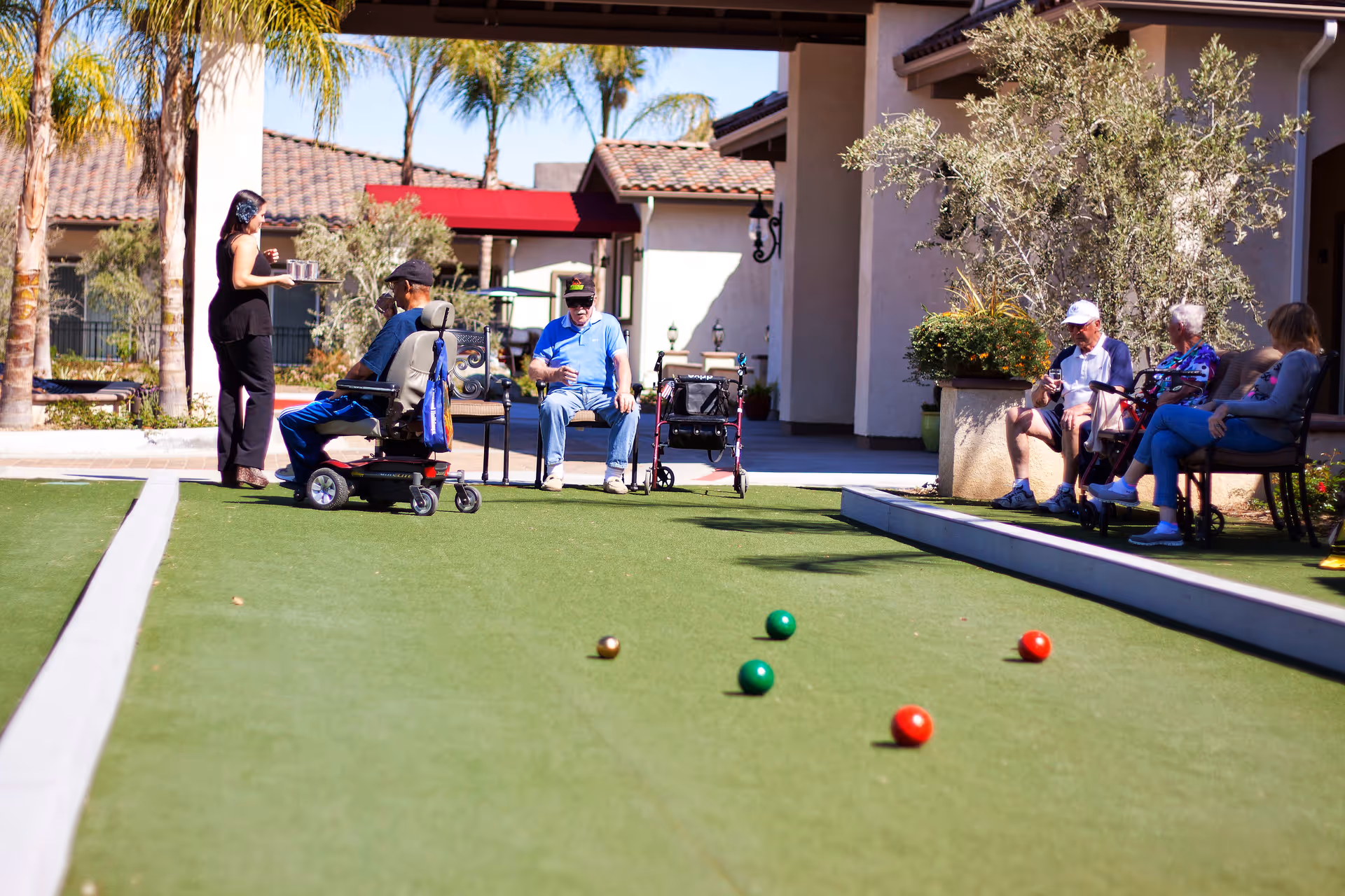 Several elderly people and a caregiver are outside on a bocce ball court at a senior living facility. Some are seated on benches and one person is in a mobility scooter. There are bocce balls on the court, and the setting includes palm trees and buildings with tiled roofs in the background.