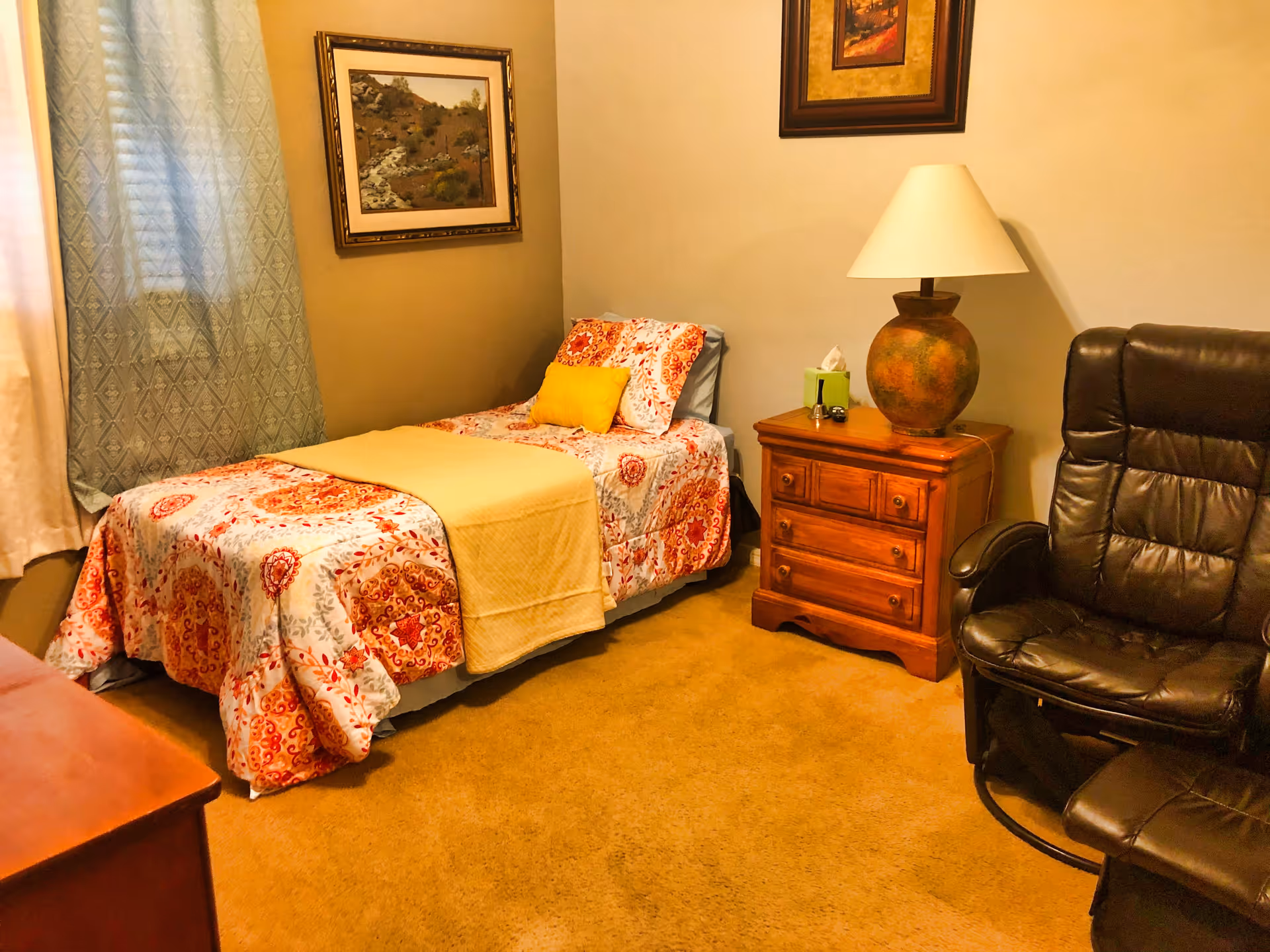 A cozy bedroom with a single bed covered in a colorful patterned bedspread and a yellow blanket. There is a wooden nightstand with a large lamp and a tissue box on it. A black leather recliner chair is positioned next to the nightstand. The room has beige walls with two framed pictures hanging and a window with patterned curtains.