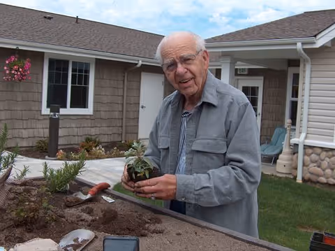 An elderly man wearing glasses and a gray jacket is gardening outdoors at a raised garden bed in a courtyard area of a senior living facility. He is holding a small plant and smiling. The background shows the exterior of the facility building with windows, doors, and some outdoor furniture.