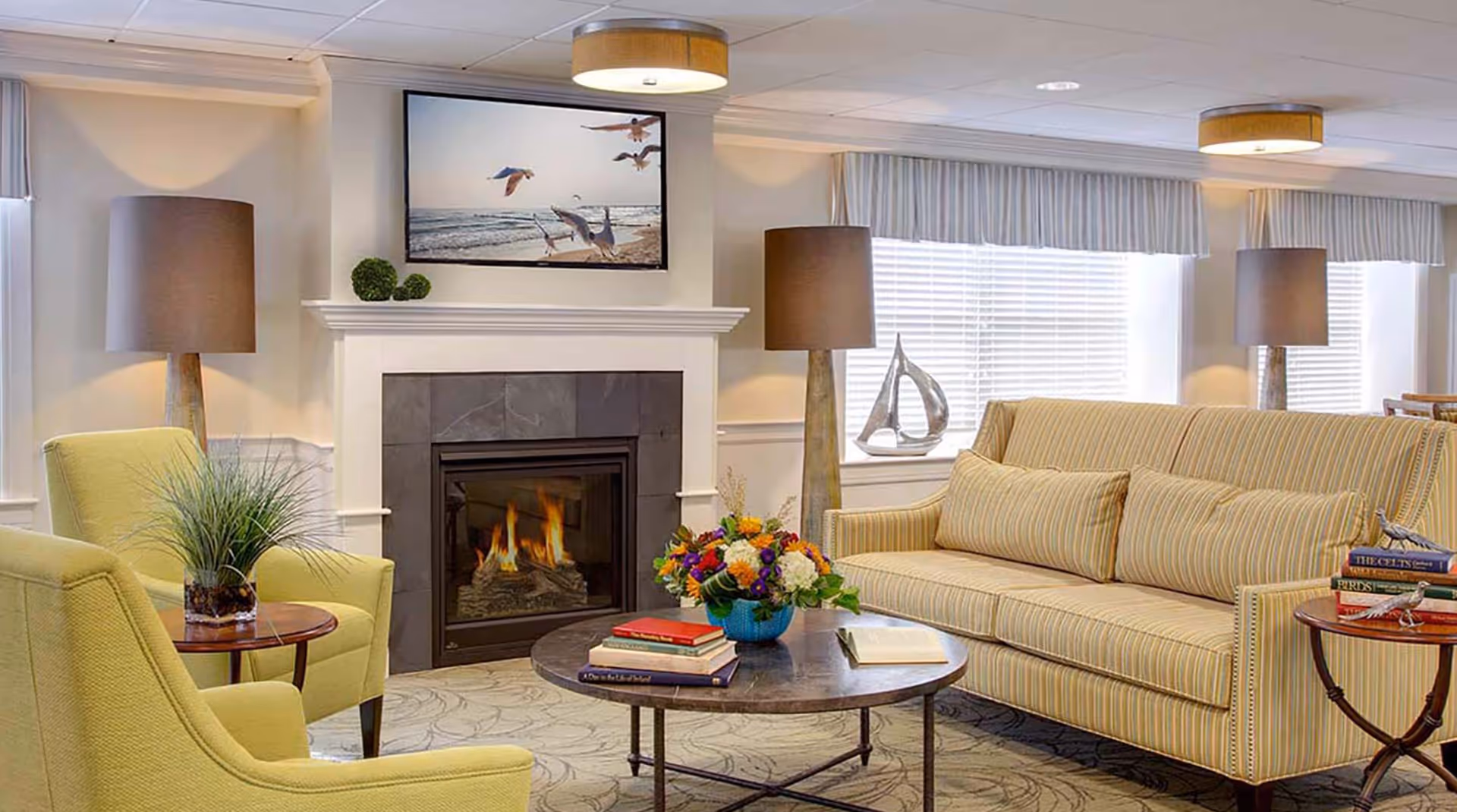 A cozy living room with a lit fireplace, a striped sofa with matching pillows, two yellow armchairs, a round coffee table with books and a flower arrangement, two side tables with books and decorative items, two tall lamps with brown shades, and a large window with blinds and valances.