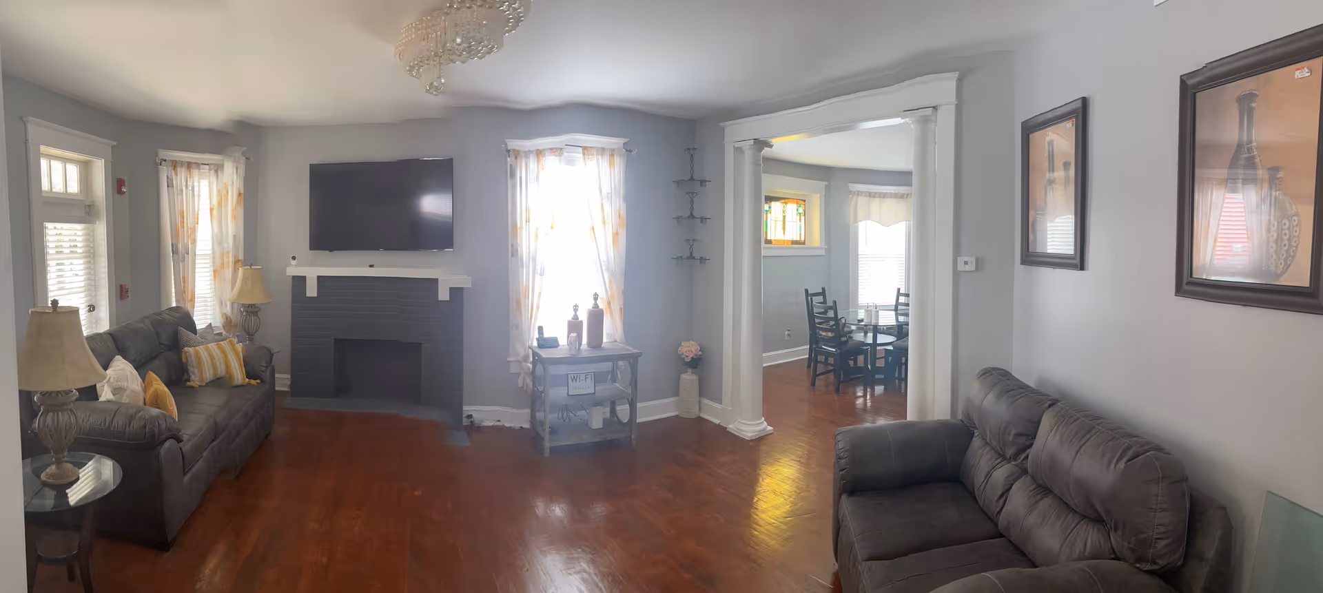A cozy living room with two gray sofas facing each other, a black fireplace with a mounted flat-screen TV above it, two windows with sheer curtains letting in natural light, a small table with decorative items and a Wi-Fi sign, and an adjacent dining area visible through a wide doorway with white columns.