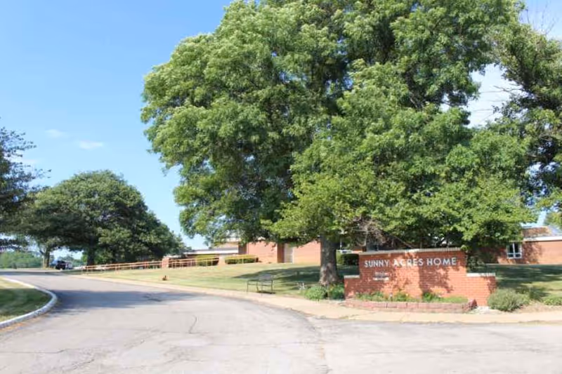 Exterior view of Sunny Acres Nursing Home showing a curved driveway, large green trees, and a brick sign with the facility name on a sunny day.