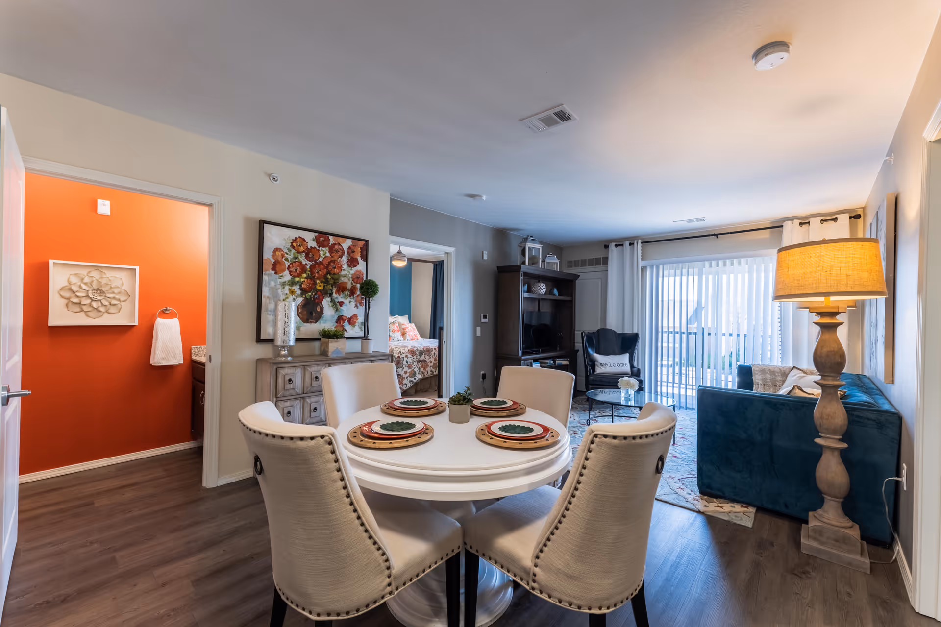 A cozy and well-lit living and dining area in a senior living facility. The dining area features a round white table set with four beige upholstered chairs and place settings. The living room has a blue sofa, a wooden floor lamp, a dark wood entertainment center with a TV, and a sliding glass door with vertical blinds leading to an outdoor space. To the left, an open door reveals a bathroom with bright orange walls and a towel hanging on the wall. Another doorway shows a bedroom with floral bedding.