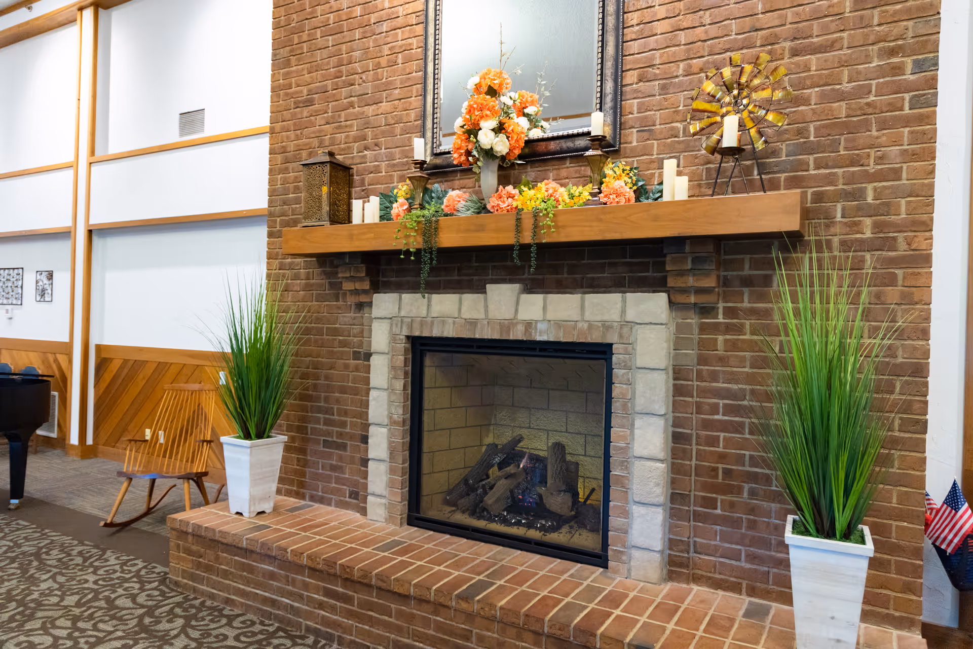 A cozy brick fireplace with a wooden mantel decorated with orange and white flowers, candles, and a small windmill ornament. Two tall green plants in white pots flank the fireplace. In the background, there is a wooden rocking chair and a piano in a well-lit room with wood paneling and carpeted floor.