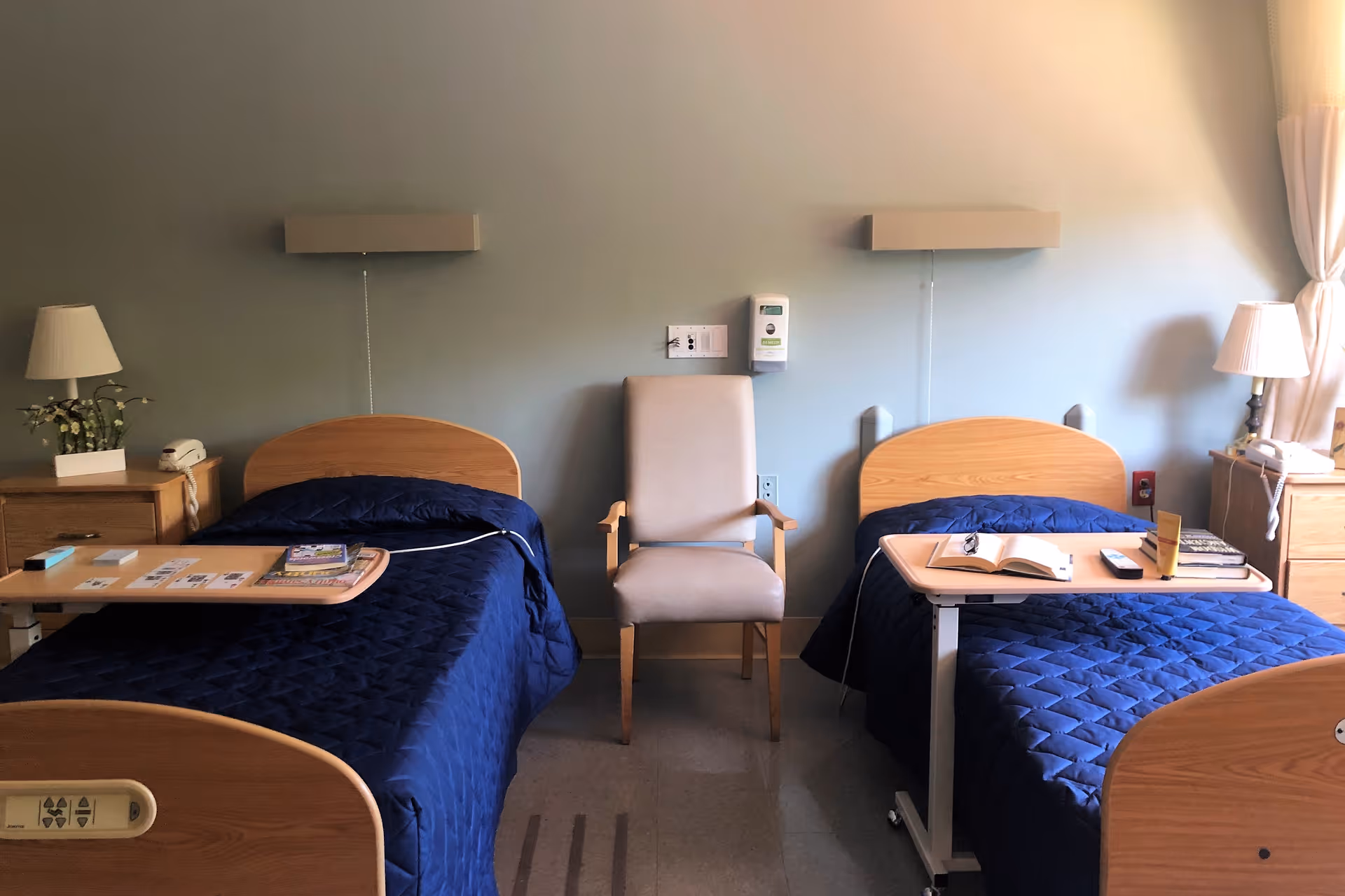 A room with two single beds covered in dark blue quilts, each with an overbed table holding books and other items. Between the beds is a beige armchair. Each side of the room has a wooden nightstand with a lamp and a telephone. The walls are light-colored with wall-mounted lights above each bed.