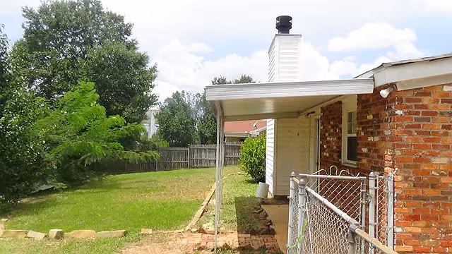 Backyard view of a residential care facility showing a grassy lawn with some trees and bushes, a wooden fence in the background, and part of a brick building with a covered patio and a chain-link gate in the foreground.