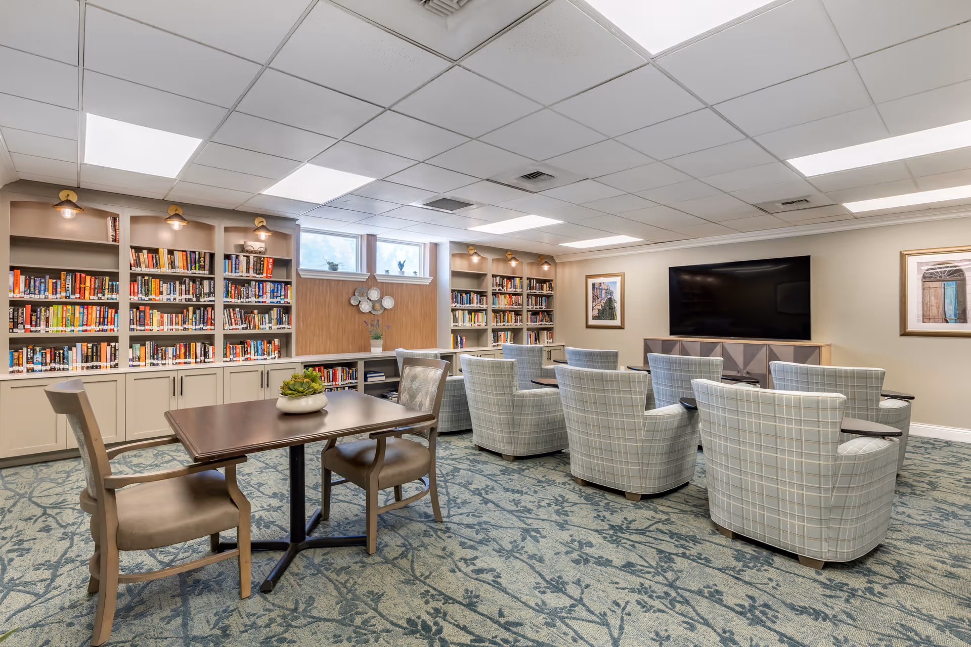 A bright communal library and media room with bookshelves along the back wall, a table and chairs in the foreground, and upholstered armchairs facing a large wall-mounted TV.