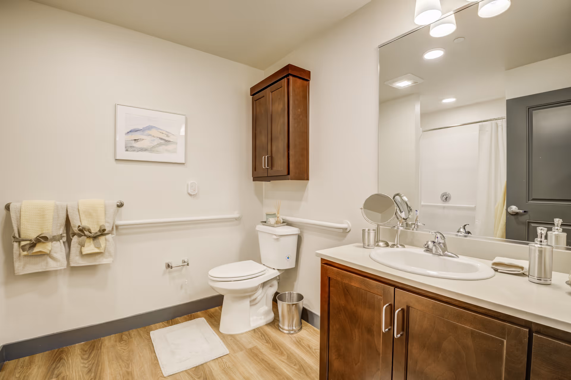 A clean and well-lit bathroom with wooden cabinets, a white toilet, a large mirror above a sink with a countertop, and a towel rack holding two beige towels. There is a small framed artwork on the wall and a white bath mat on the wooden floor.