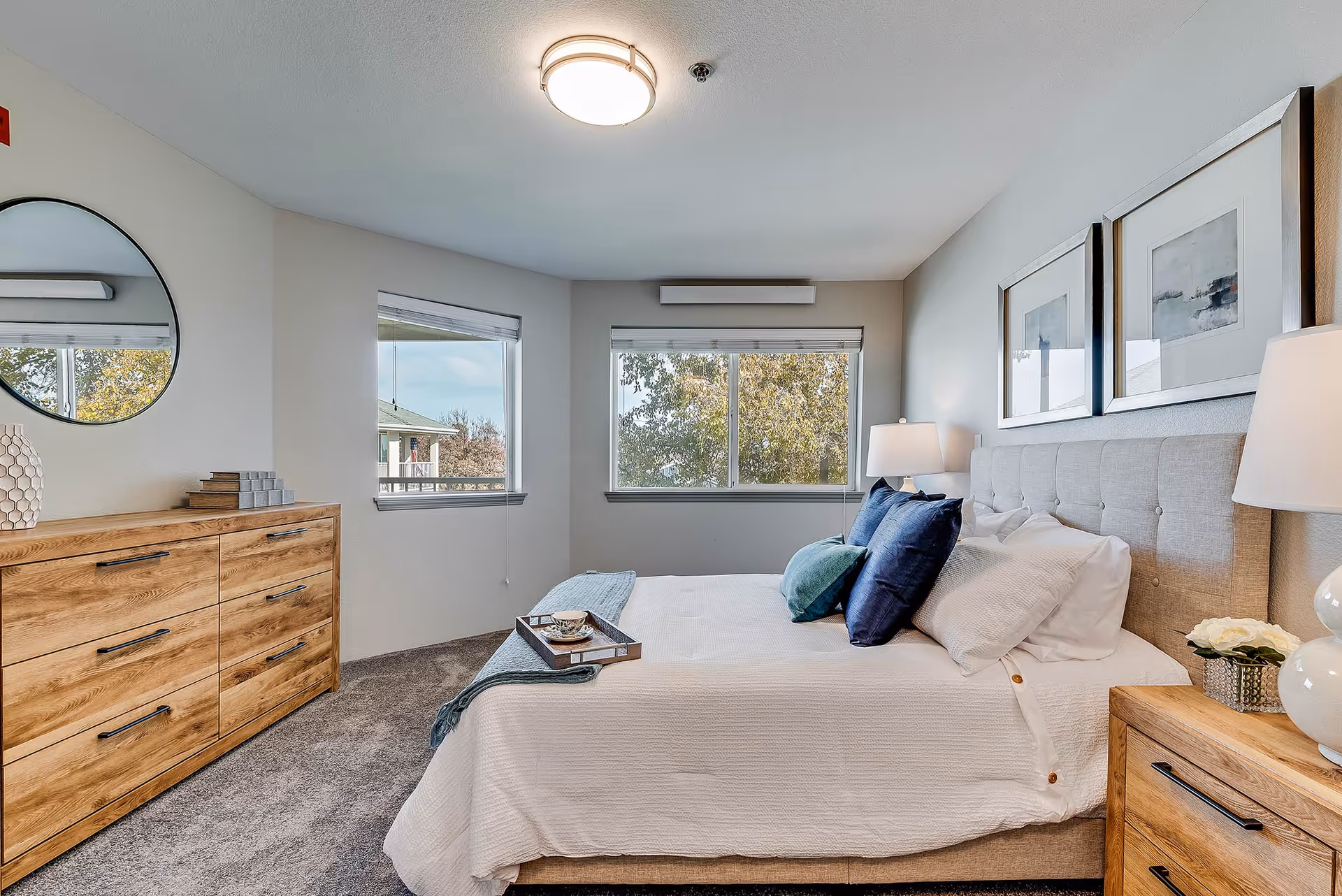A bright and cozy bedroom with a large bed featuring white bedding and blue accent pillows. The room has two windows with views of trees outside, a wooden dresser with a round mirror above it, and two framed artworks on the wall above the bed. There are bedside tables with lamps and decorative items on each side of the bed.