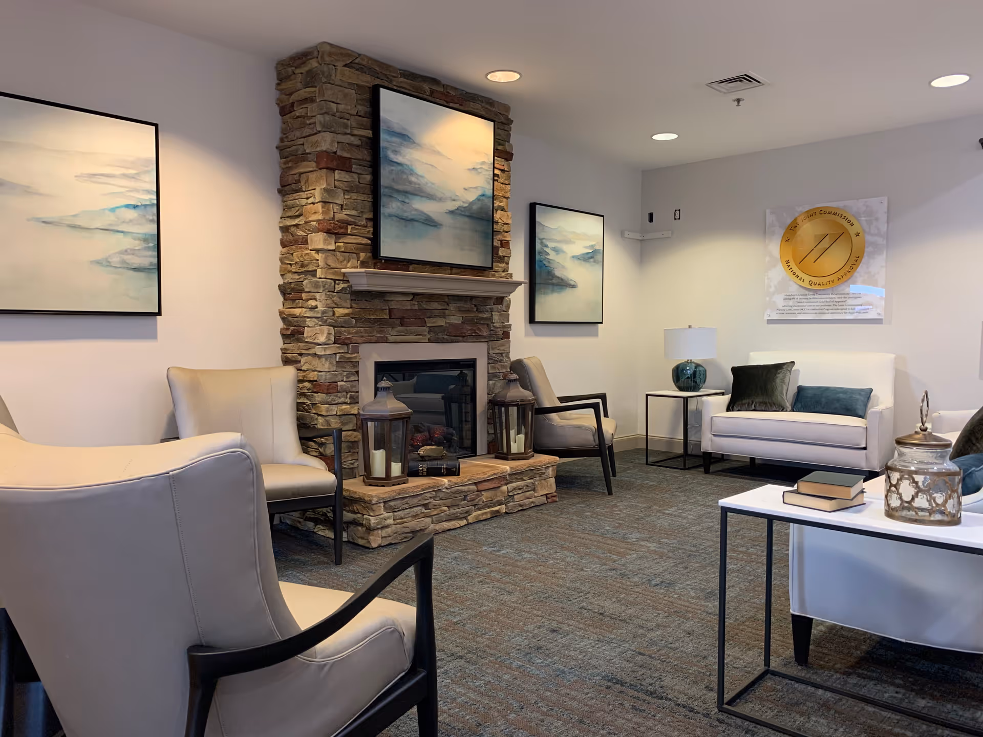 A cozy living room area with a stone fireplace in the center, flanked by two beige armchairs. Above the fireplace is a framed abstract painting with blue and white tones. On the walls are two additional similar paintings. To the right, there is a white loveseat with dark green and blue cushions, next to a small side table with a lamp. In the foreground, a white coffee table holds a decorative jar and a couple of books. The room has a carpeted floor and recessed ceiling lights.
