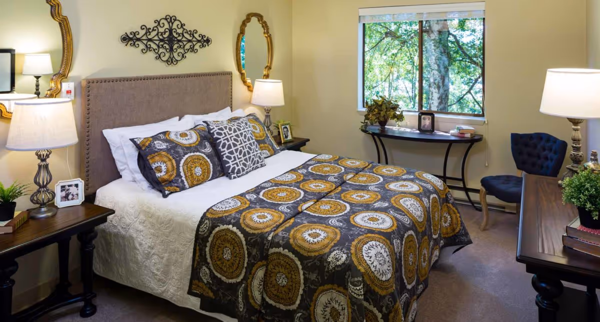 A cozy bedroom in an assisted living community featuring a neatly made bed with a patterned comforter and multiple pillows. The room includes two bedside tables with lamps, decorative mirrors on the wall, a window with a view of green trees, a small table with plants and framed photos, and a blue upholstered chair.