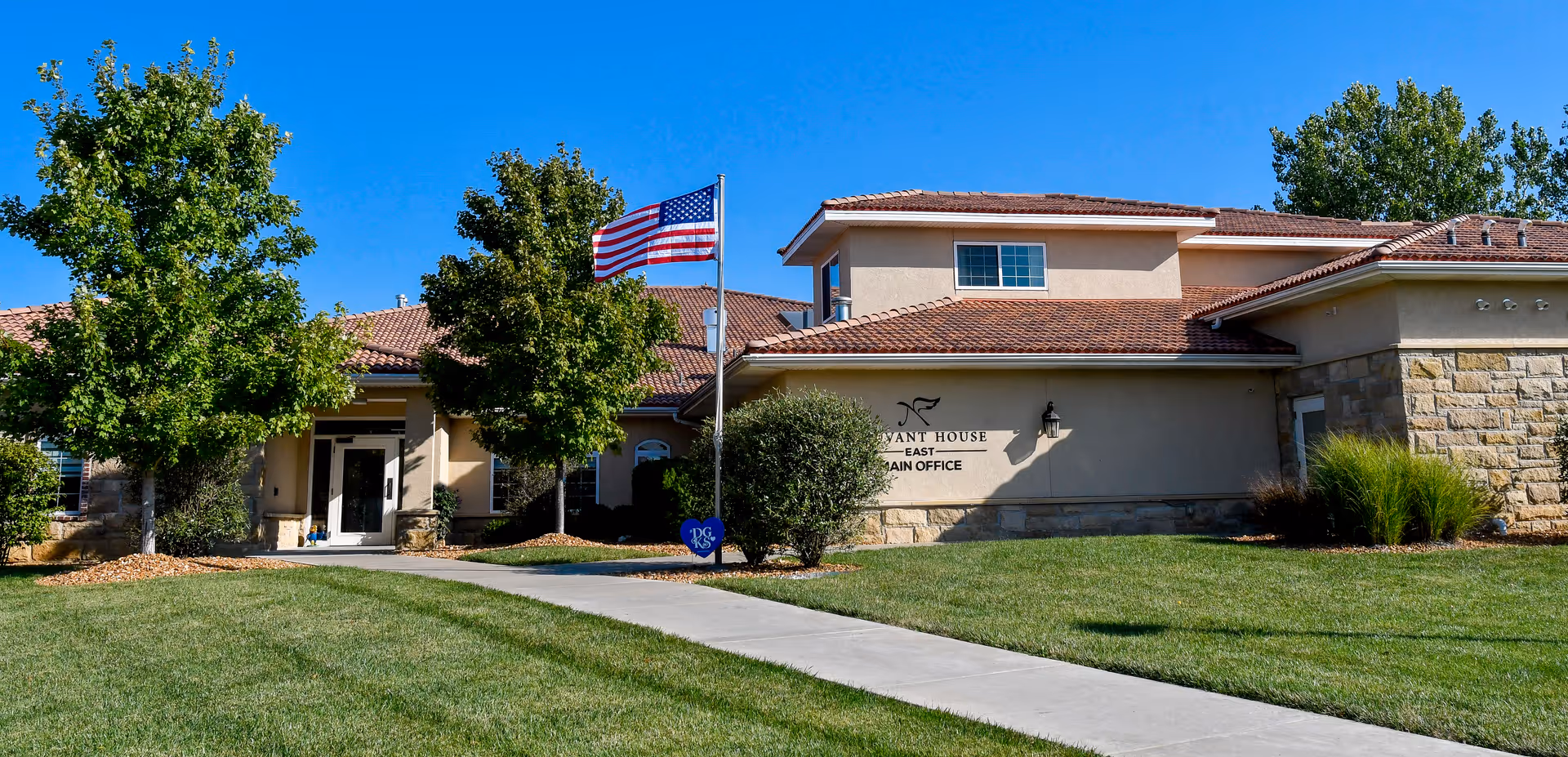 Exterior view of Neuvant House of Lawrence building with a well-maintained lawn, a concrete walkway, two trees, an American flag on a flagpole, and clear blue sky.